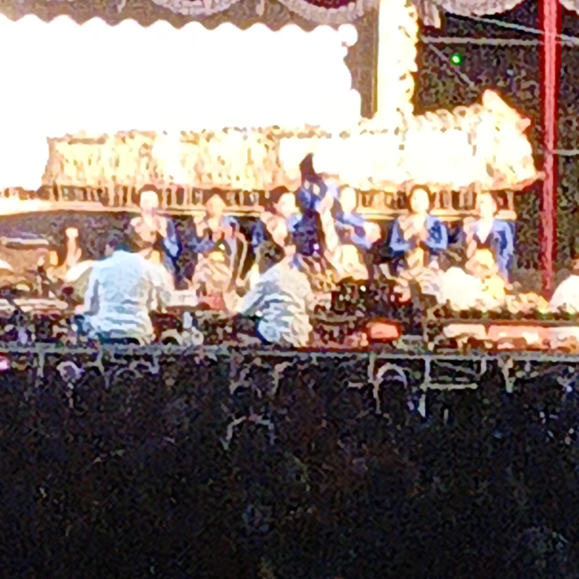 The image depicts a performance in a dimly lit outdoor setting, likely a stage with a decorative canopy overhead. In the foreground, a dark, textured surface suggests a tiered seating area or a stage platform. Behind this, several individuals are seated, some appearing to be musicians. One person in the center, viewed from the back, wears a white shirt and is seated, possibly playing an instrument. Other figures are dressed in blue garments. A golden, ornate structure, possibly part of a musical instrument or a decorative element, is visible on the left. A small, bright green light is present in the upper right quadrant of the image. The overall impression is of a cultural performance, potentially part of a festival or event in Mangli, Indonesia.
