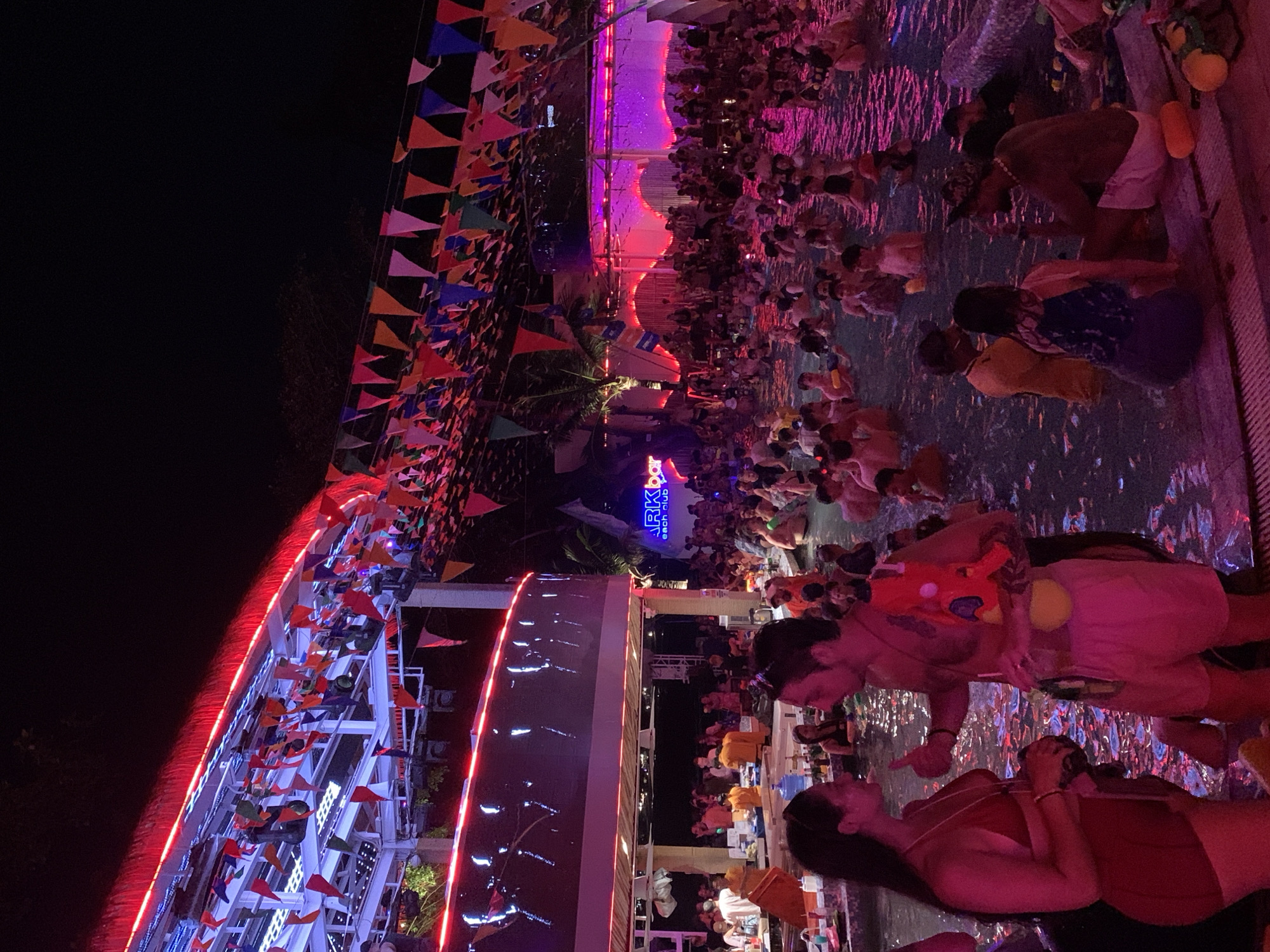 The image depicts a crowded pool party at Ark Bar Beach Club in บ้านบางรักษ์, Thailand. The scene is at night, illuminated with colorful lights and decorations. Numerous people are in the pool, some holding water guns. There are pennant banners strung across the scene. A multi-level bar is adjacent to the pool.