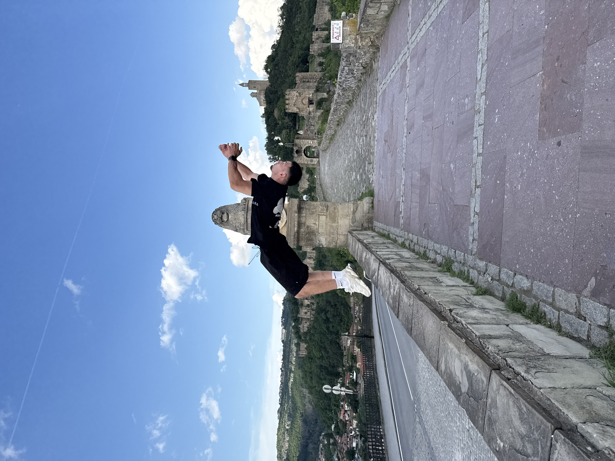 A man is captured mid-air in a jump, appearing to defy gravity, at a high vantage point in Veliko Tarnovo, Bulgaria. The man is of Asian descent, wearing a black t-shirt, black shorts, and white athletic shoes. He is positioned over the edge of a stone structure. His arms are raised in front of him, seemingly in a celebratory gesture.
Behind the man is a panoramic view of Veliko Tarnovo. It reveals buildings nestled amongst a green, hilly landscape. The cloudy blue sky is visible above. Part of the Tsarevets Fortress is visible on a hill in the background. The immediate foreground consists of the stone structure on which the man is standing, showing a pathway of reddish stone tiles. A road lies directly beneath the stone edge.