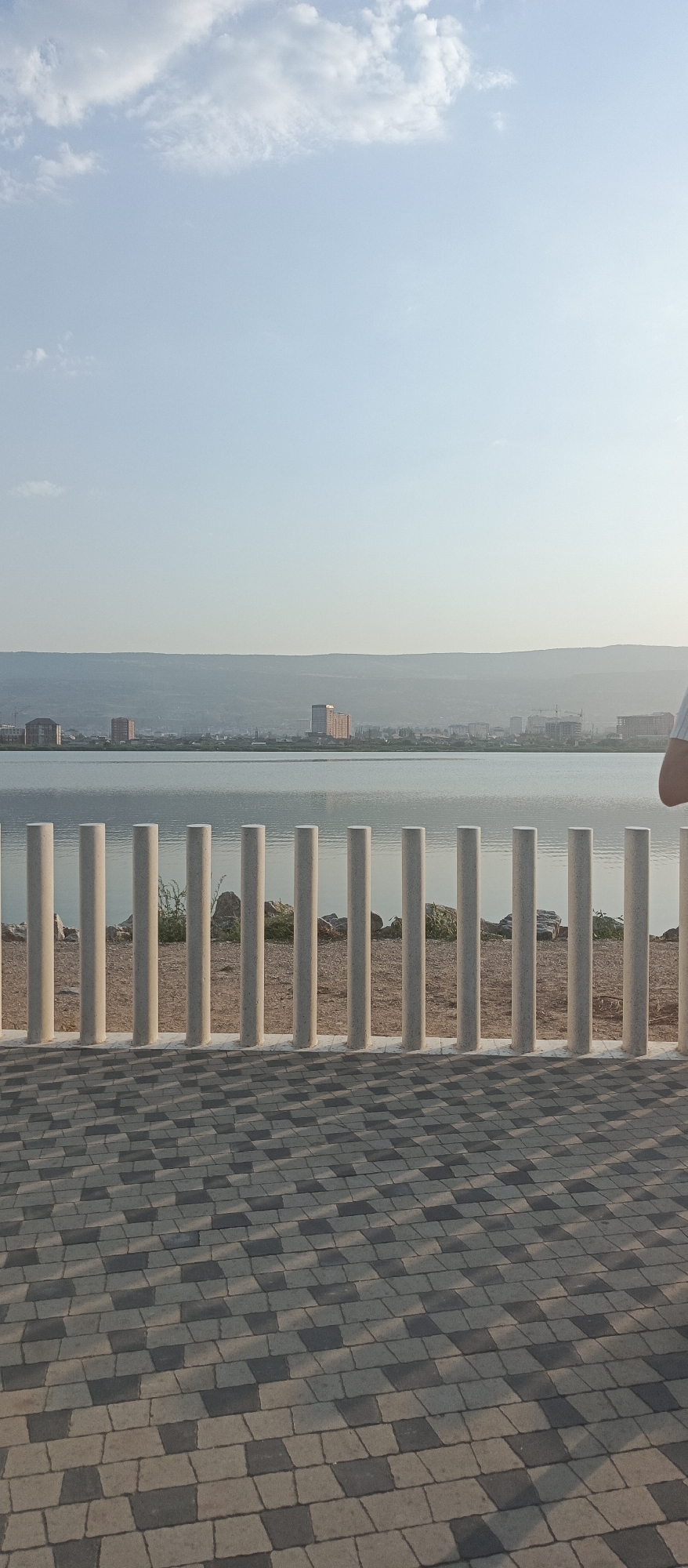 A vertical, eye-level shot captures a waterfront promenade in Makhachkala, Russia. In the foreground, a patterned pavement of grey and black rectangular tiles leads the eye toward a row of evenly spaced, light-colored cylindrical posts that form a barrier. Beyond the posts, a tranquil body of water reflects the clear, pale blue sky, with scattered wispy clouds. Along the distant shoreline, the city of Makhachkala is visible, featuring various buildings, including several tall, modern structures. Mountains form a hazy backdrop to the cityscape. A person's shoulder and arm, wearing a white t-shirt, are visible on the right edge of the frame, suggesting they are standing at the promenade, facing the water. The overall scene is one of peaceful urban waterfront scenery.