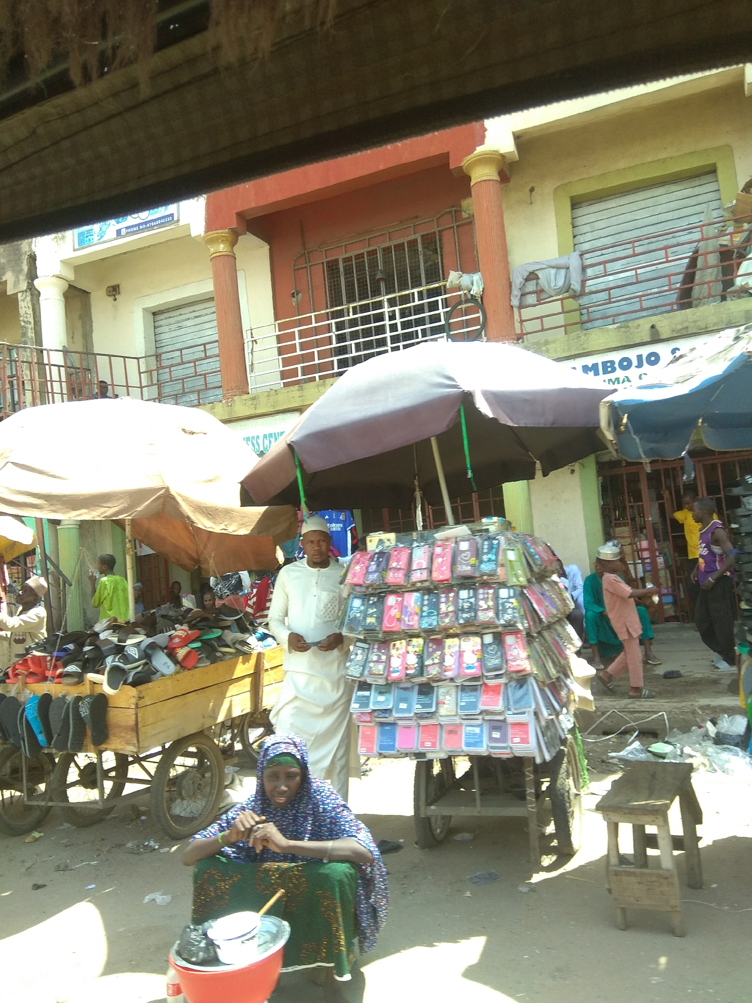 In Jalingo, Nigeria, a bustling street market scene unfolds under a bright, possibly midday sun. The foreground is occupied by a woman in a patterned headscarf and a long, colorful garment, seated on the ground behind a red basin containing a white bowl and other items. She appears to be tending to her wares.
To her right, a wooden cart filled with an assortment of footwear, primarily sandals and flip-flops in various colors, is positioned. Dominating the midground is a stall featuring a large display of mobile phone cases, stacked in multiple rows and presented on a tiered structure attached to a cart. A man in a light-colored, traditional outfit stands near this display, seemingly attending to customers or overseeing the stall. Behind him, other individuals are visible, including a young boy in a green garment.
In the background, a two-story building with red and yellow accents lines the street. The building features windows with metal grates and shutters, some of which are partially open. Signage is visible on the building, with one sign displaying "PHONES NO. 07066642333" and another partially obscured sign reading "MBОЈО". The street itself is unpaved, typical of many urban areas in Nigeria, with scattered debris and evidence of foot traffic. Two large umbrellas, one beige and one dark gray, provide shade over parts of the market. The overall impression is one of active commerce and daily life in a Nigerian city.