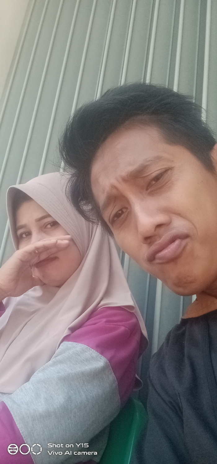 A selfie-style image depicts two individuals, one male and one female, positioned in front of a light greenish-grey background featuring distinct vertical ridges. The male, partially cropped on the right side of the frame and closer to the camera, has dark hair and is wearing a dark-colored top. He exhibits a pouting facial expression with furrowed eyebrows and pursed lips. The female, positioned behind and to the left of the male, wears a light beige hijab and a long-sleeved top with a magenta body and grey lower sleeves. Her right hand is raised, partially covering her mouth and nose. Her visible left eye is directed towards the camera. A fragment of a green object, possibly a chair, is visible in the lower right background. The image bears a "Shot on Y15 Vivo AI camera" watermark at the bottom left. No specific visual cues within the image confirm the location as Gending, Indonesia.