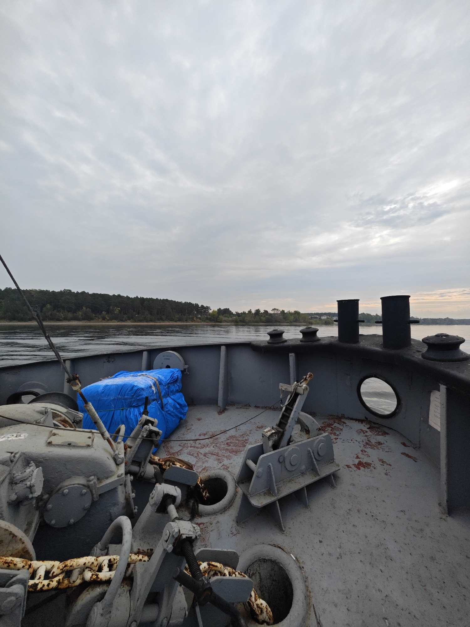 This image depicts the deck of a vessel, likely a barge or tugboat, on a cloudy day. The foreground is dominated by ship equipment, including a large, rusty anchor chain coiled and attached to a winch mechanism. A bright blue tarp-covered object is secured on the deck. In the midground, the vessel's deck continues with bollards and a circular porthole. The background shows a calm body of water, a tree-lined shore with some scattered buildings, and a hazy sky. The location is identified as Ермолаево, Russia. There are no people visible in the image.