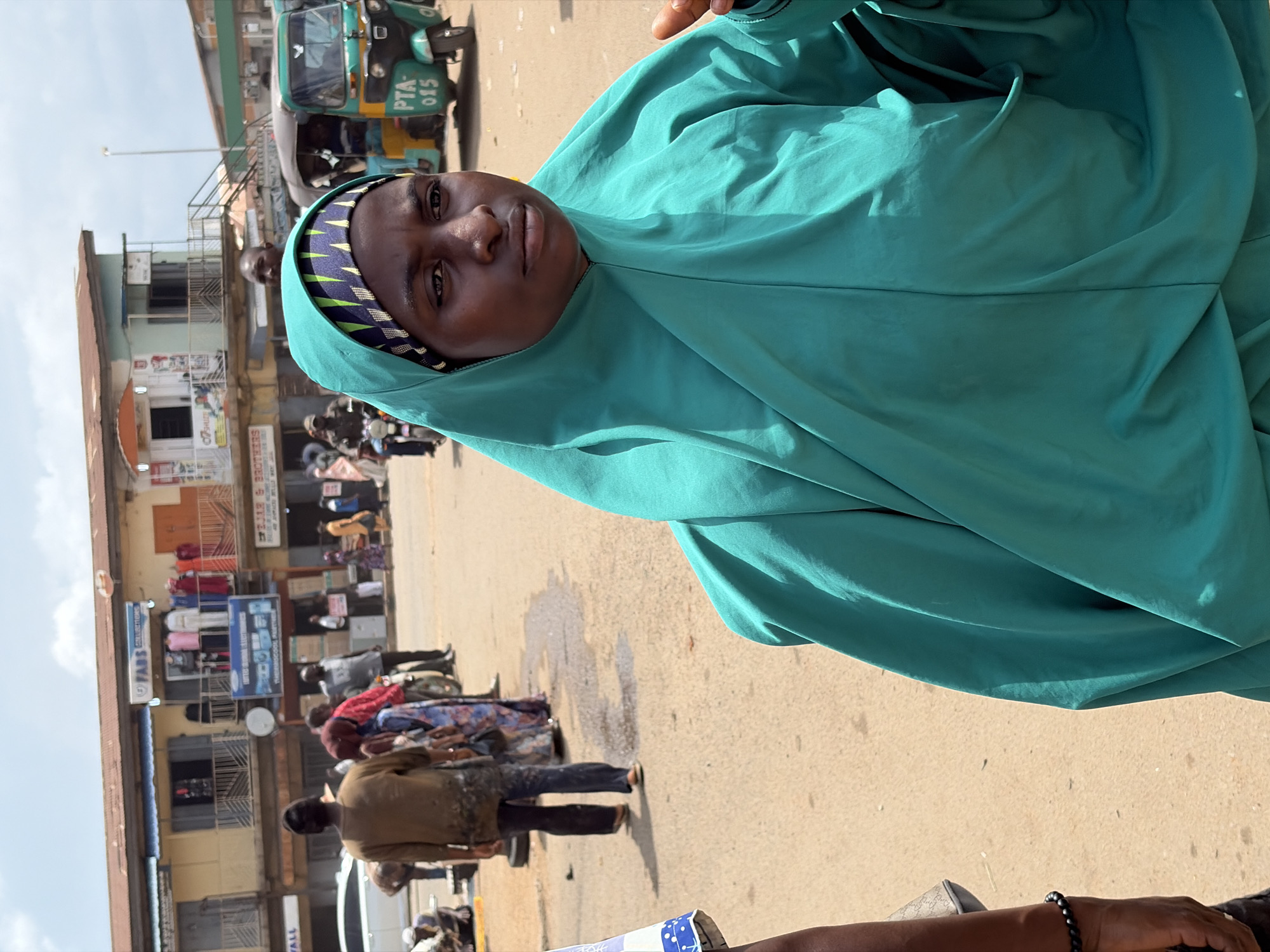 The image depicts an outdoor scene in a commercial area of Jos, Nigeria. In the foreground, a woman wearing a teal-colored hijab and headscarf is the primary subject. She is looking to her right with a neutral expression. Behind her, a busy street is visible with several people, vehicles, and buildings. A green and yellow auto-rickshaw, identified by the license plate "PTA 015", is parked on the right side of the street. Several shops with signage like "FABS COLLECTIONS" and "BROTHERS" line the street. People are seen walking and standing around, some carrying bags. A white car is parked on the left side of the street. The ground is a dusty, paved surface, with some wet patches suggesting recent moisture. The sky above is overcast with clouds. The overall impression is of a typical day in a bustling market or commercial district.
