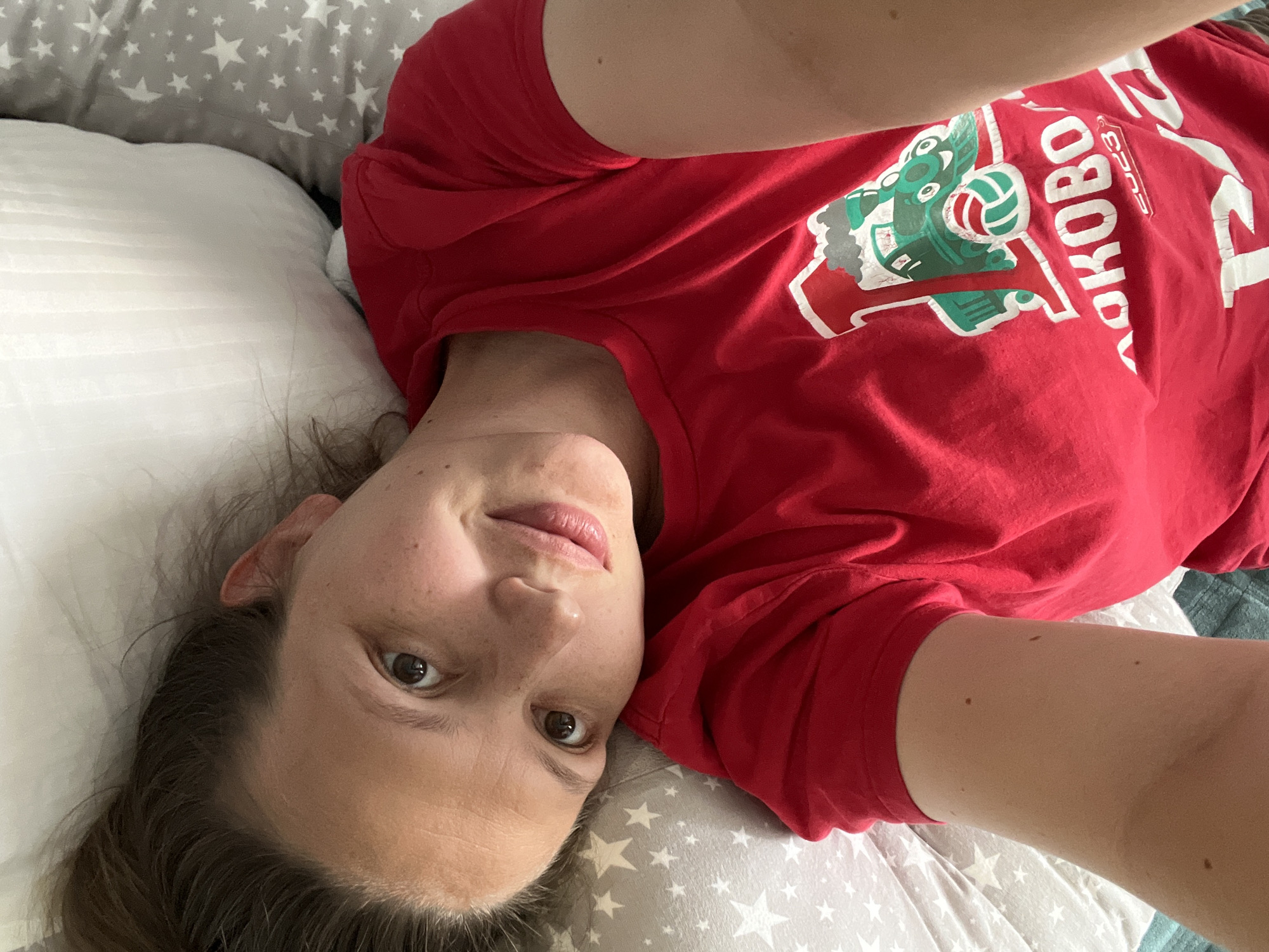 The image features a woman lying on her back, looking directly at the camera with a neutral expression, possibly with a slight smile. Her head is positioned towards the left of the frame. She wears a red t-shirt with a green graphic design depicting a mascot-like figure holding a volleyball, accompanied by Russian text including "ЛОКОВО" (likely part of "Локомотив") and the year "2023." Her dark brown hair is spread around her head on a white, striped pillow. A grey pillow with a white star pattern is visible behind her head. A blue-green blanket or sheet is partially visible on the right side of the frame. The scene is indoors, likely a bedroom, illuminated by natural light, suggesting it is daytime. The location is Chaikovsky, Russia.