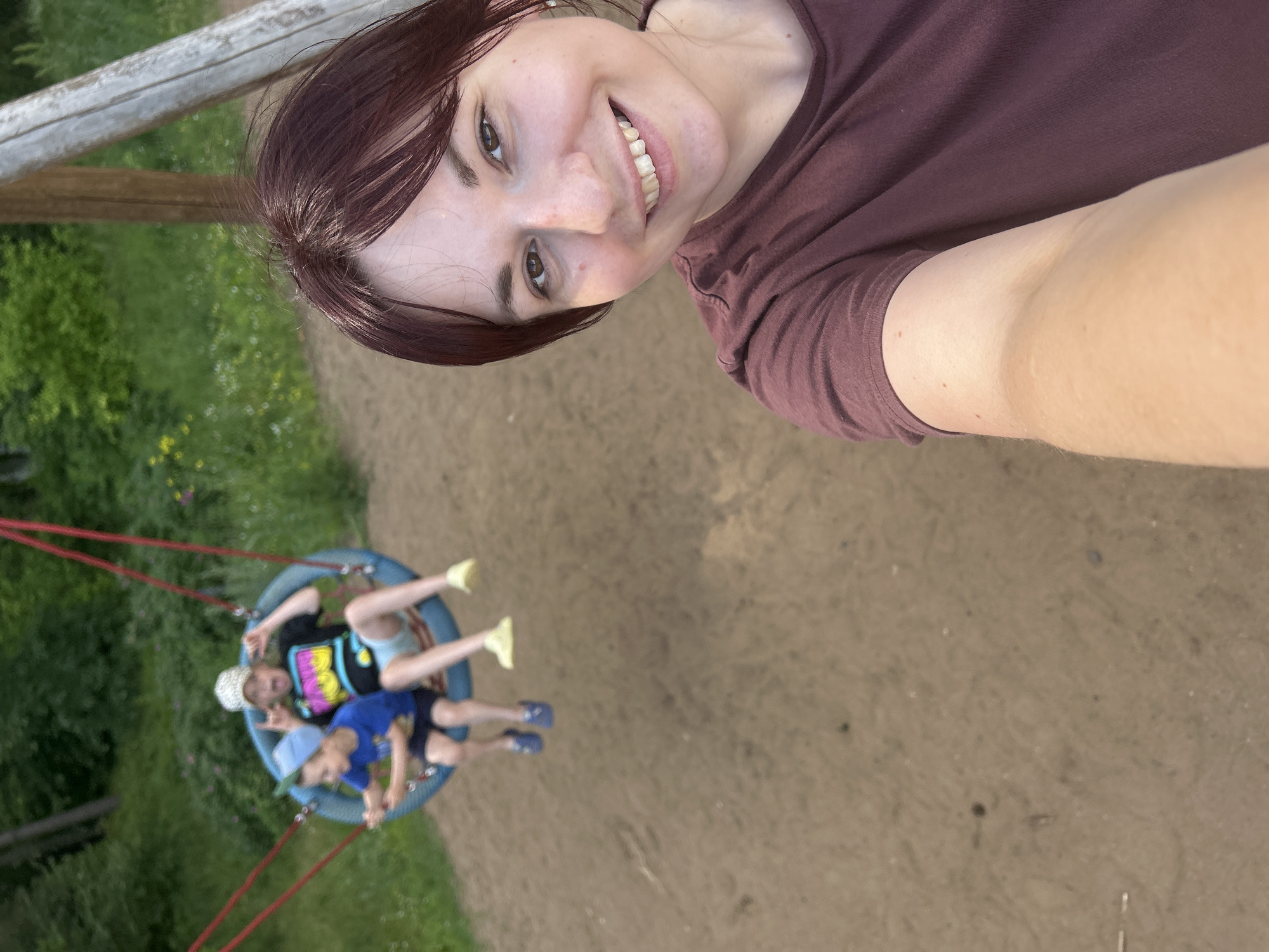 In a park setting with sand and greenery, a woman with dark hair and a maroon t-shirt takes a selfie. Behind her, two children are on a tire swing. The child on the left wears a blue t-shirt and blue shorts. The child on the right wears a black t-shirt with a colorful design, a white patterned cap, and light-colored shorts. The swing is suspended by red ropes from a wooden structure. The background shows trees and foliage, indicative of an outdoor location. This scene is situated in Chaikovsky, Russia.