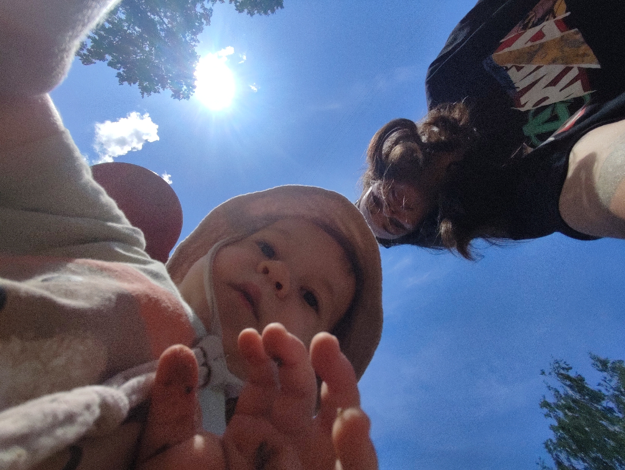 The image shows a low-angle, first-person perspective of a baby in a tan sun hat reaching out with their hands. The baby's face is visible, with dark eyes and a neutral expression. Behind the baby, a person's torso and head are visible, wearing a black t-shirt with a graphic print. The background is a bright blue sky with a shining sun and some tree branches and leaves. The scene appears to be outdoors on a sunny day. There is no specific indication of the location within the city of Chaikovsky, Russia.