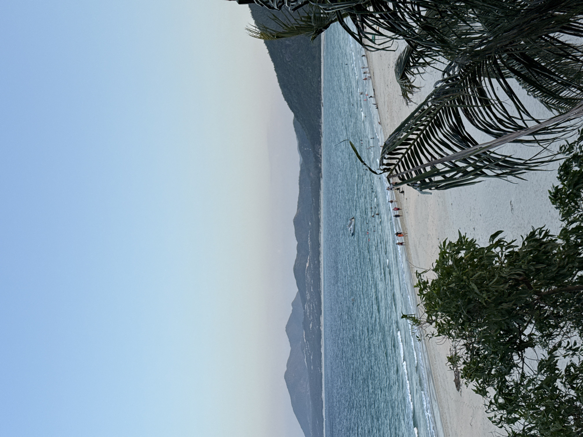 The image depicts a serene beach scene in Song Cau, Vietnam, likely during late afternoon or early evening. The sky is a soft gradient of pale blue, transitioning to a lighter hue near the horizon. In the distance, a range of lush, green mountains forms the backdrop, their peaks softened by the atmospheric haze.
The turquoise ocean stretches out, with gentle waves lapping onto the sandy shore. A white motorboat is visible in the water, and several people can be seen enjoying the beach, some walking along the shoreline and others in the water.
In the foreground, the leaves of a palm tree are prominently featured, their dark fronds creating a natural frame for the scene. To the left, the foliage of another tree adds a touch of vibrant green to the composition. The sand is light-colored, and there are subtle textures and shadows indicating the presence of footprints and small debris on the beach. The overall impression is one of a tranquil coastal landscape.