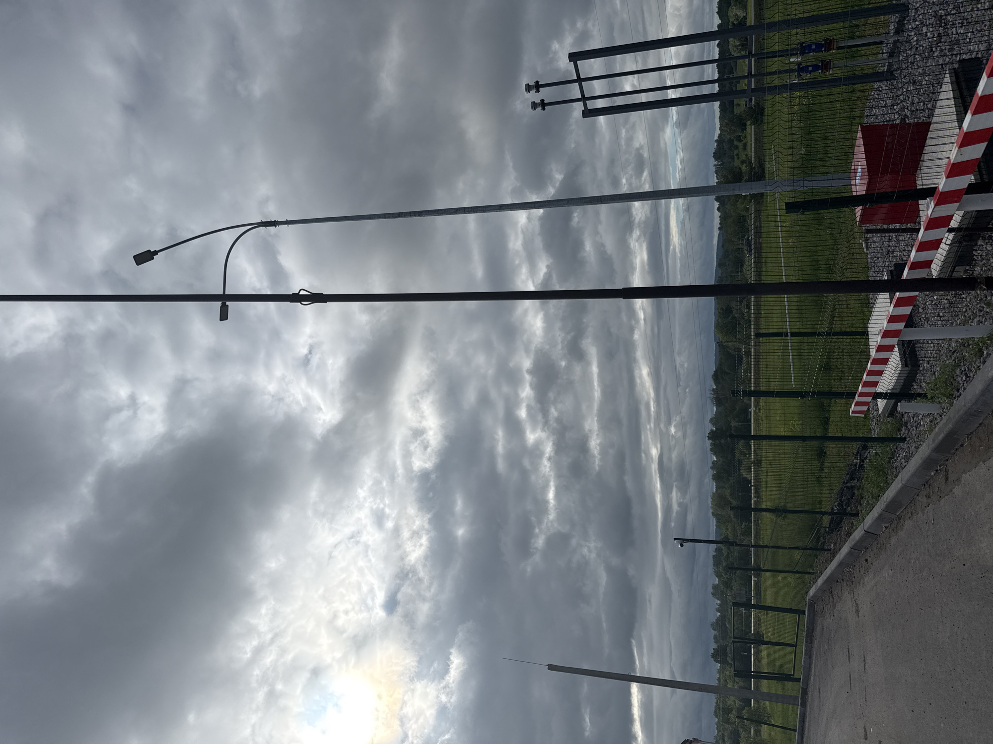 The image captures an outdoor scene under a dramatic, heavily clouded sky, with shades of grey dominating the upper and central portions. Patches of brighter light, suggesting the sun, are visible breaking through the clouds towards the lower left. The foreground features a paved path or road with a concrete curb running horizontally across the bottom.
Several tall, slender utility poles or light posts are prominent subjects, extending vertically across the frame and appearing silhouetted against the sky. One significant pole in the mid-left has two curved lampposts attached. Further to the right, a security fence, likely made of metal mesh, separates the paved area from an expansive green field. A red and white striped barrier post is visible on the right, positioned next to a red rectangular utility box. Additional metal structures, including one with horizontal bars and insulators, are seen near the fence line on the right. Small, dark security cameras (CCTVs) are mounted on some of the poles and fence posts, indicating surveillance.
In the background, beyond the green field, a dense line of trees or forest stretches along the horizon, with hints of distant, undulating terrain. No people or specific activities are depicted, giving the scene a quiet, almost contemplative atmosphere. The heavy cloud cover suggests an overcast day, possibly late afternoon or approaching evening. There is no discernible text visible in the image. The overall impression is that of a secured or infrastructure-heavy area set within a natural, rural or semi-rural landscape in Гремяча, Russia.