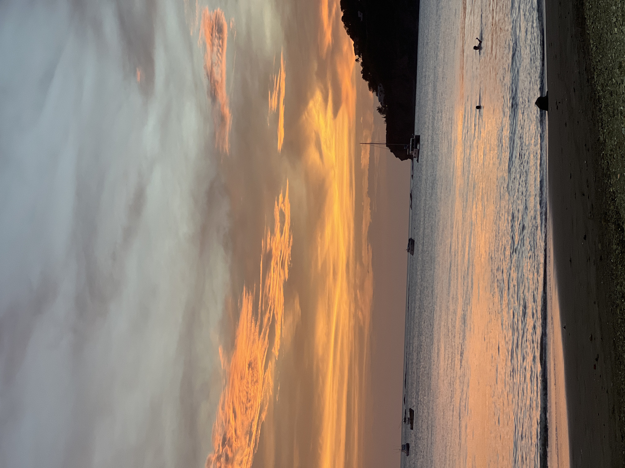 A serene sunset casts a warm orange and pink glow across the sky and the calm waters of San Juan del Sur, Nicaragua. The beach in the foreground is dark sand with small pebbles. Several boats are visible in the distance, some anchored and others moving. A few people are in the water, one appearing to swim. A dark, tree-covered cliff face rises on the right side of the image, with a few buildings visible at its summit. The overall scene is peaceful and picturesque, typical of a tropical evening.