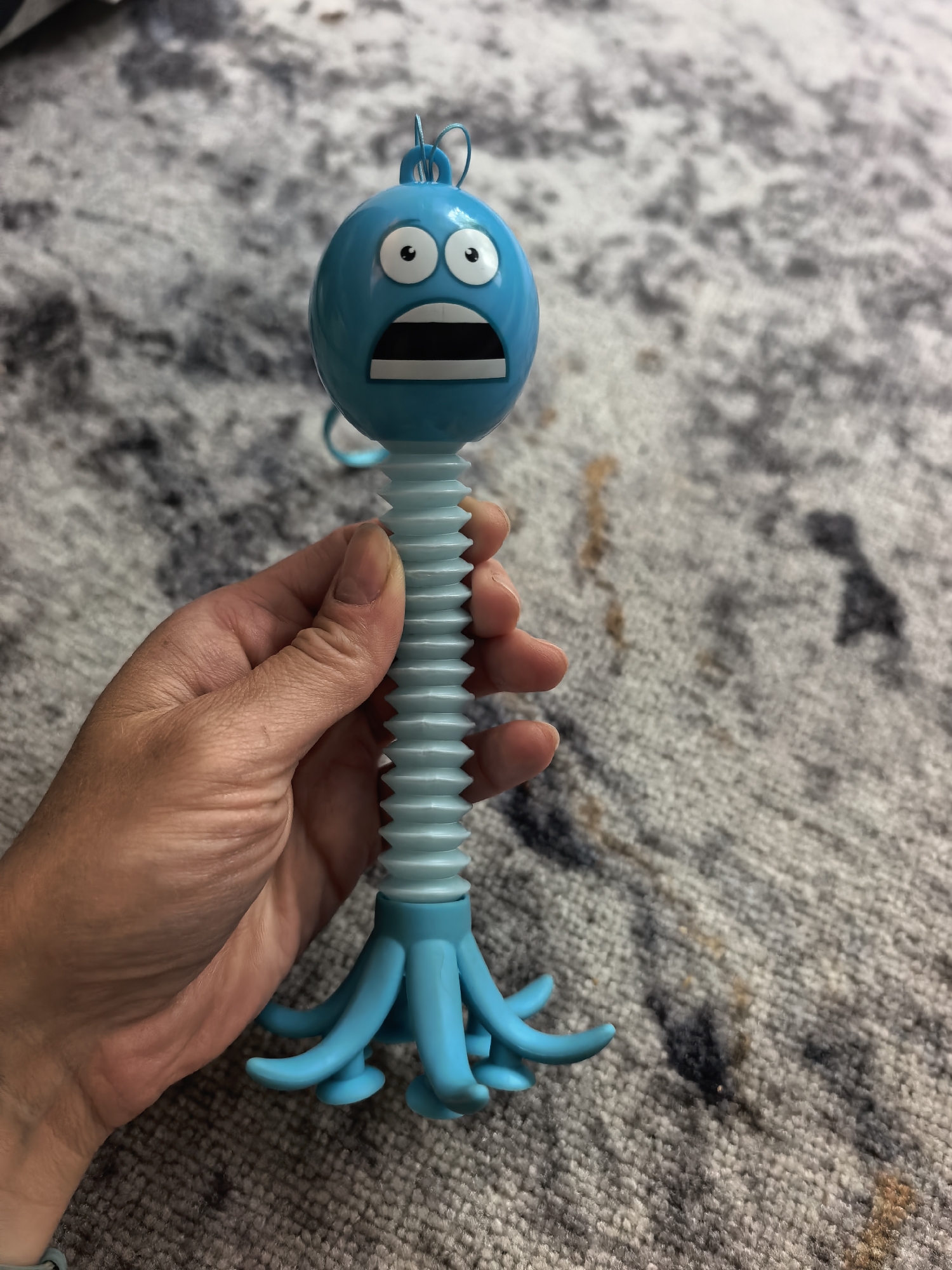 A hand holds a blue, plastic, toy character with a round head, large, surprised eyes, and an open mouth. The toy has a corrugated neck and a base of tentacles. The background is a gray carpet with a mottled pattern. The location is undetermined, and no information suggests the scene is in Sterlitamak, Russia.