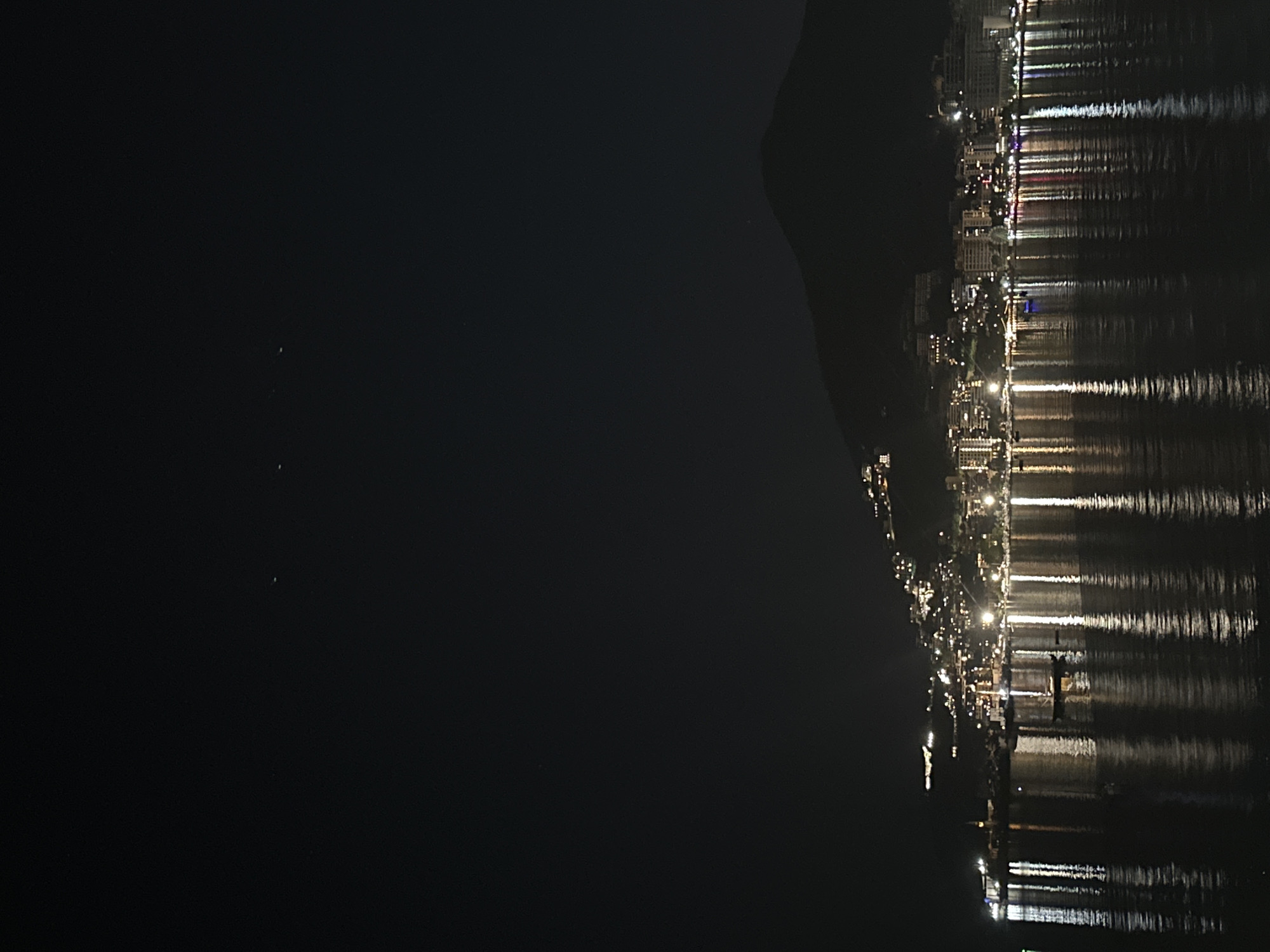 The image shows a night view of Budva, Montenegro, specifically the coastal area. The right side of the image features a brightly lit shoreline with buildings reflected in the dark water. A dark, elevated landmass is visible between the water and sky. The left side of the image is predominantly dark, suggesting the open sea.