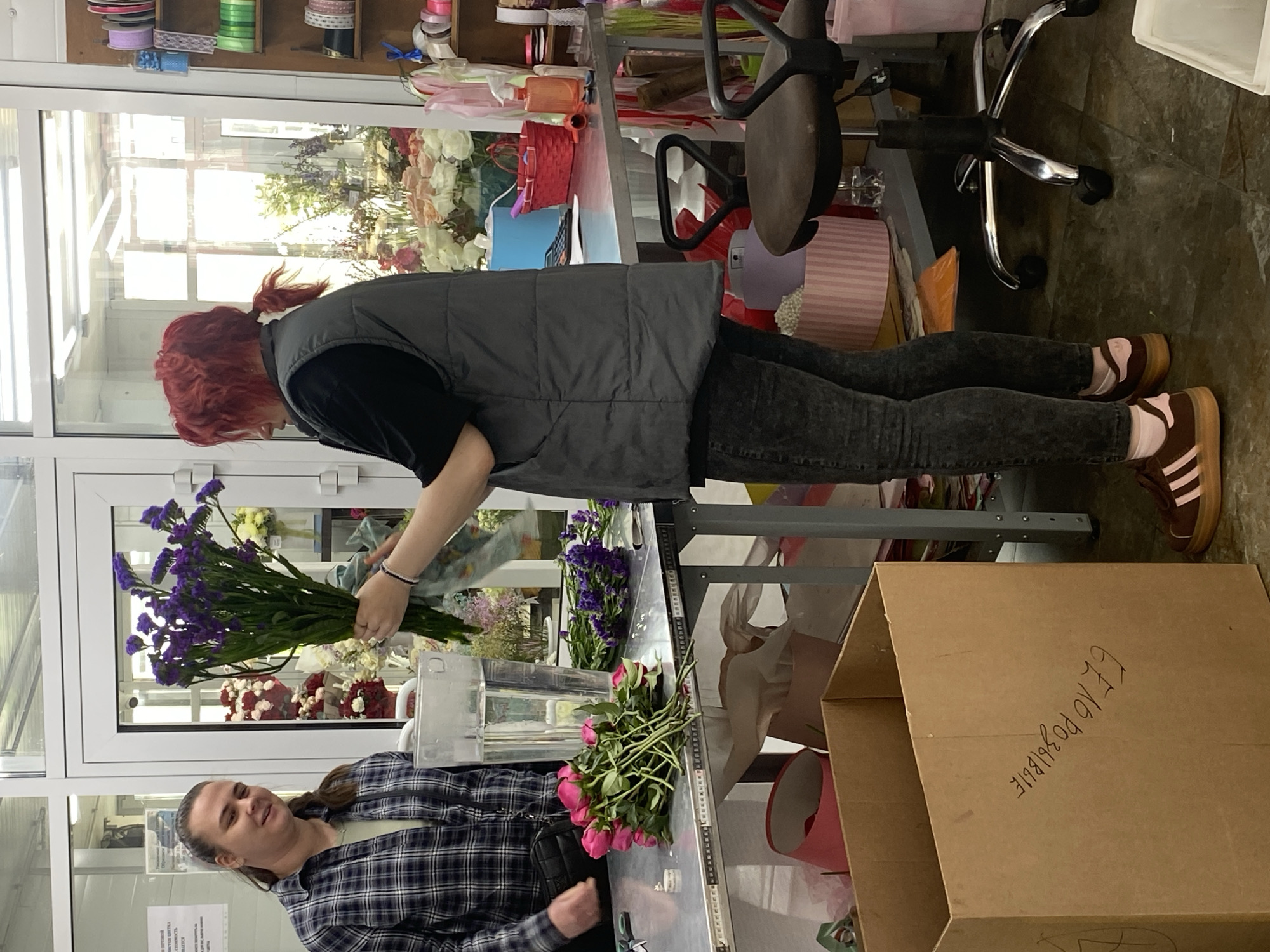 Here is a description of the image:
The image captures a scene inside what appears to be a flower shop. Two women are present. One woman, with red hair and wearing a grey vest over a black shirt and dark jeans, is arranging a bouquet of purple flowers. The other woman, wearing a blue and black plaid shirt, is near the table where pink flowers are also present. They are standing next to a metal table holding flowers, a clear container of water, and florist tools. To the side of the table is a large cardboard box with handwritten text in Russian, "Е1 по Розалие." The environment includes various flowers, ribbons, and florist supplies. The scene is illuminated by natural light, possibly from a large window. The location is identified as Yekaterinburg, Russia. A rolling office chair is visible in the background.