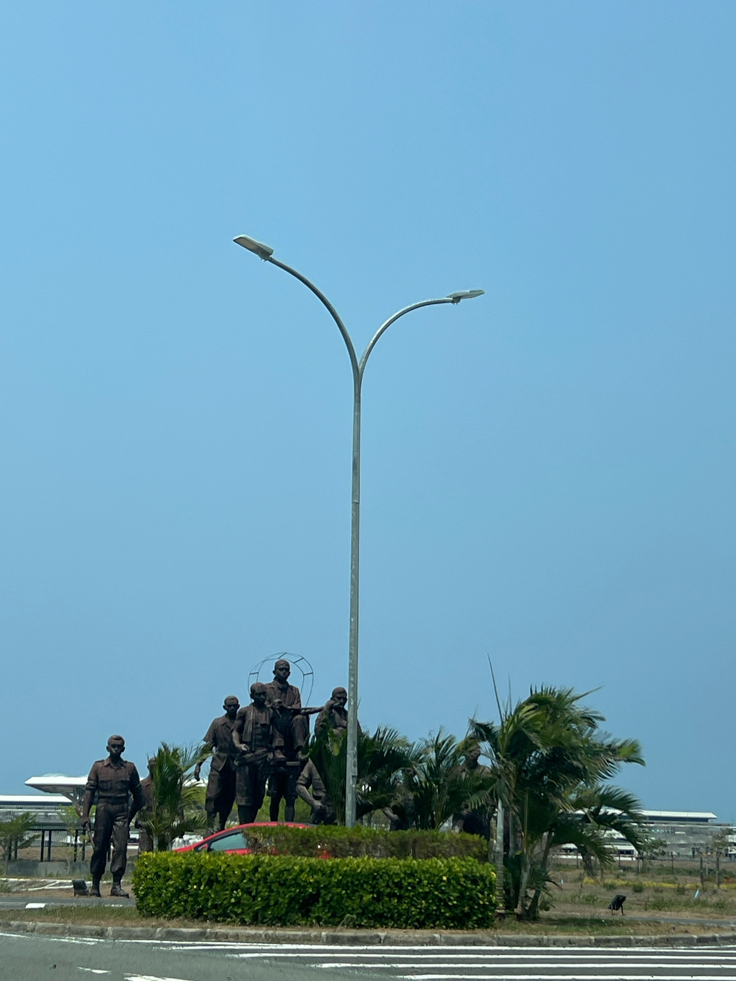 A monumental bronze statue, depicting a group of men, stands in an open area, possibly a park or roundabout, in Glagah, Indonesia. The statue is situated behind a manicured green hedge and next to palm trees. A tall, modern streetlamp with a distinctive curved design arches over the statue. In the background, the expansive blue sky is visible, with hints of industrial or institutional buildings in the distance. A red car is partially obscured by the hedge. The foreground shows a paved road with white markings. The overall scene suggests a public space, likely a monument commemorating a historical event or figures.