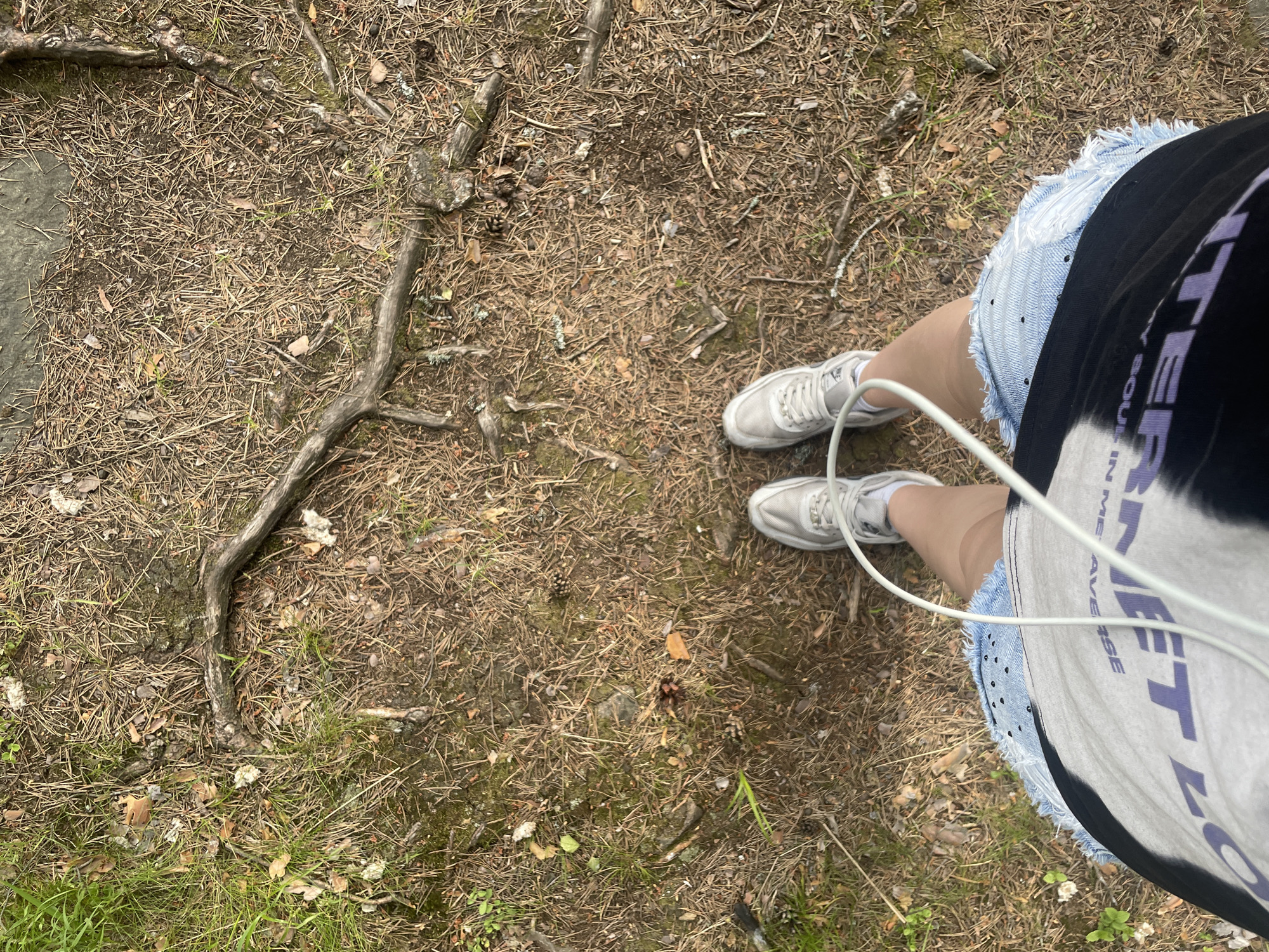 This image, captured in Boitsovo, Russia, presents a downward-facing perspective of a person standing on a forest floor. The ground is extensively covered by dry, brown pine needles, interspersed with patches of green grass, small green plants, and scattered twigs, leaves, and pinecones. Several dark, gnarled tree roots are visible traversing the surface. The lower torso, legs, and feet of the individual are depicted. They are wearing light-colored athletic shoes with white socks, light blue distressed denim shorts adorned with small dark polka dots, and a black and white/grey tie-dye t-shirt featuring partial purple text "TER" and smaller, lighter text "MY SOUL IN ME AVER". White earbud wires descend from above the frame, passing in front and between the person's legs. Small, dark, cap-like objects, consistent with mushrooms, are visible in clusters on the ground in several areas.