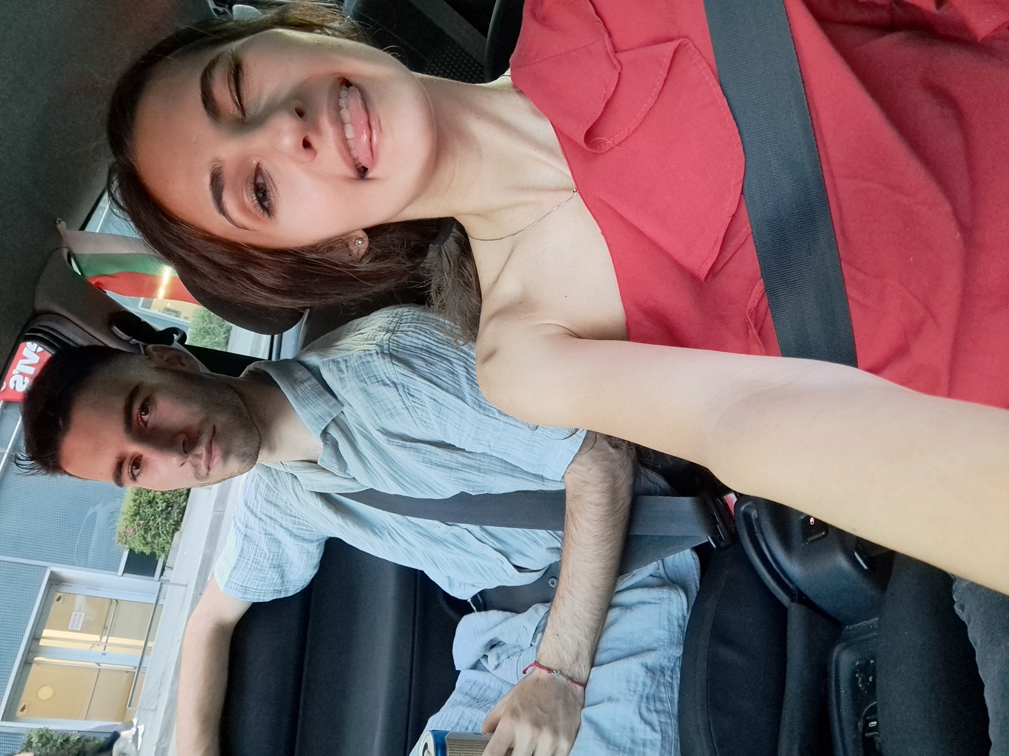 Two individuals are inside a vehicle, seemingly a car. A young woman with dark hair and a red shirt is in the passenger seat, sticking her tongue out playfully. She is wearing a seatbelt. A male individual is in the driver's seat, wearing a light blue shirt, also with his seatbelt fastened. A Bulgarian flag is visible hanging from the car window. The car is parked in front of a building with glass doors and windows. Based on the Bulgarian flag, it can be inferred that the location is likely in Sofia, Bulgaria.