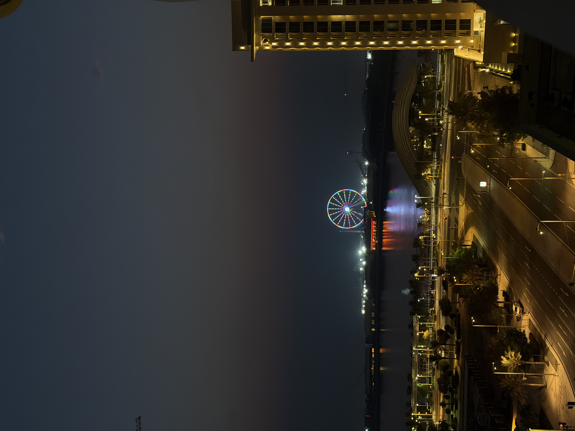 The image depicts a nighttime view of Lusail, Qatar, dominated by a large, illuminated Ferris wheel in the distance. The Ferris wheel features a vibrant display of colorful lights, standing out against the dark sky. In the foreground, a tall, modern building with illuminated windows is visible on the right side of the frame. A body of water reflects the lights of the city and the Ferris wheel. The middle ground shows a waterfront area with structures and more lights, as well as a road with streetlights illuminating the pavement and some palm trees. A single white car is parked on the side of the road. The overall scene captures the urban landscape of Lusail at night, characterized by its bright lights and modern architecture.