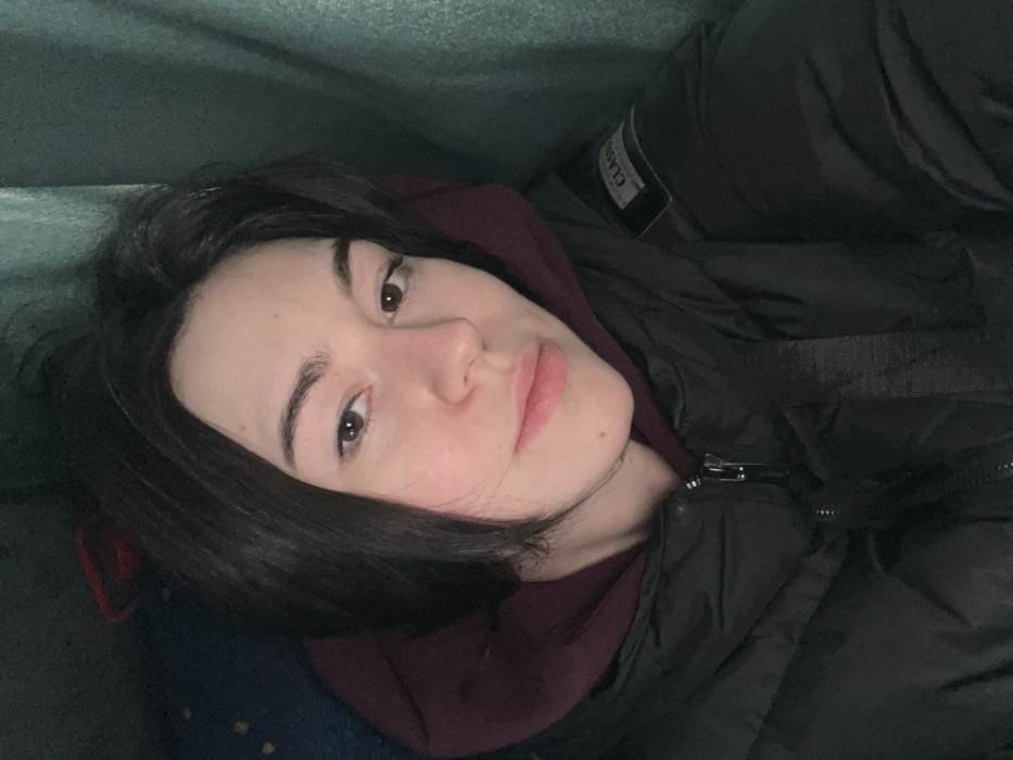 The image depicts a female individual, likely a young adult, captured in a close-up portrait within an interior setting in the city of Hatne, Ukraine. She has dark brown, short-to-medium length hair, parted on the left, and fair skin. Her brown eyes are directed towards the camera, and her lips are slightly parted. A small mole is visible below her lower lip on the right side. She is wearing a dark, possibly black, puffer jacket over a dark red or maroon hooded sweatshirt, the hood of which is visible around her neck. A black fabric strap, possibly from a bag or a seatbelt, crosses diagonally over her chest. A square patch with the word "CLASSIC" and other smaller text is visible on the upper right arm of her jacket. The background on the left consists of dark blue patterned fabric, while the background on the right features a dark teal or green fabric, suggesting upholstery or curtains within an enclosed space, such as a vehicle.