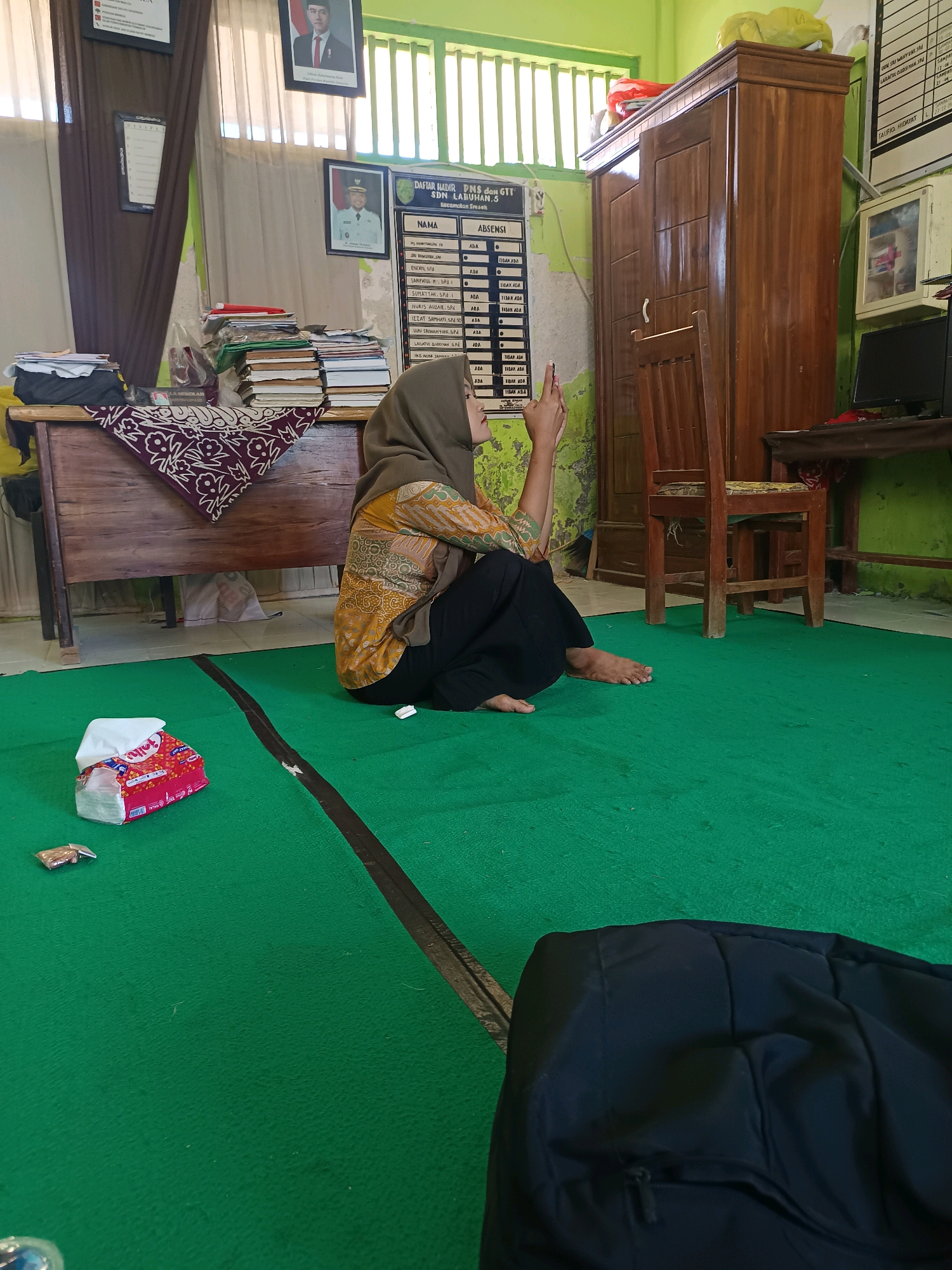 The image depicts an indoor scene, likely a classroom or office, in Labuhan, Indonesia. A young woman wearing a hijab and a patterned blouse is seated on the floor, which is covered with a green carpet. She is looking towards the right side of the frame with her left hand raised, possibly holding a phone. In the foreground, a packet of tissues and a small metallic object are on the carpet. To the left, a wooden desk is laden with books and papers. Further in the background, a tall wooden cabinet stands against a green wall. There are framed pictures and a bulletin board displaying a list of names and attendance statuses on the wall. Sunlight streams in from a window with vertical blinds on the upper left side of the image.