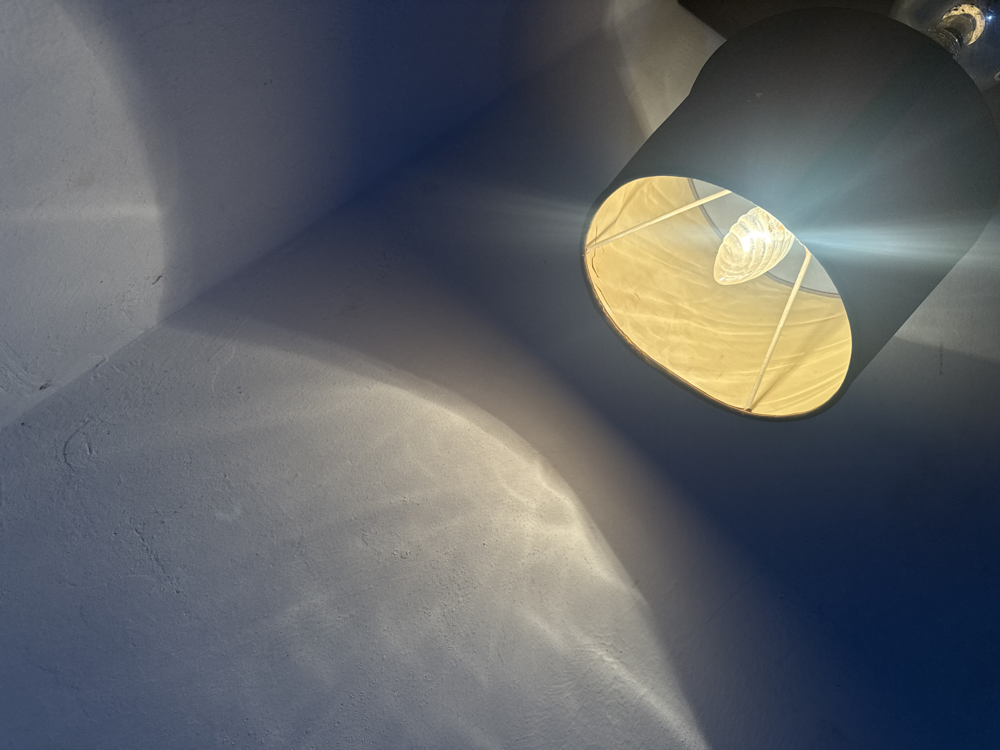 A room is illuminated by a lamp with a black lampshade and a visible lightbulb inside, mounted on the ceiling. The light casts an oval-shaped illumination on the white-painted textured wall, with a shadow directly below the lamp. The location of the image is not possible to determine based on the picture.