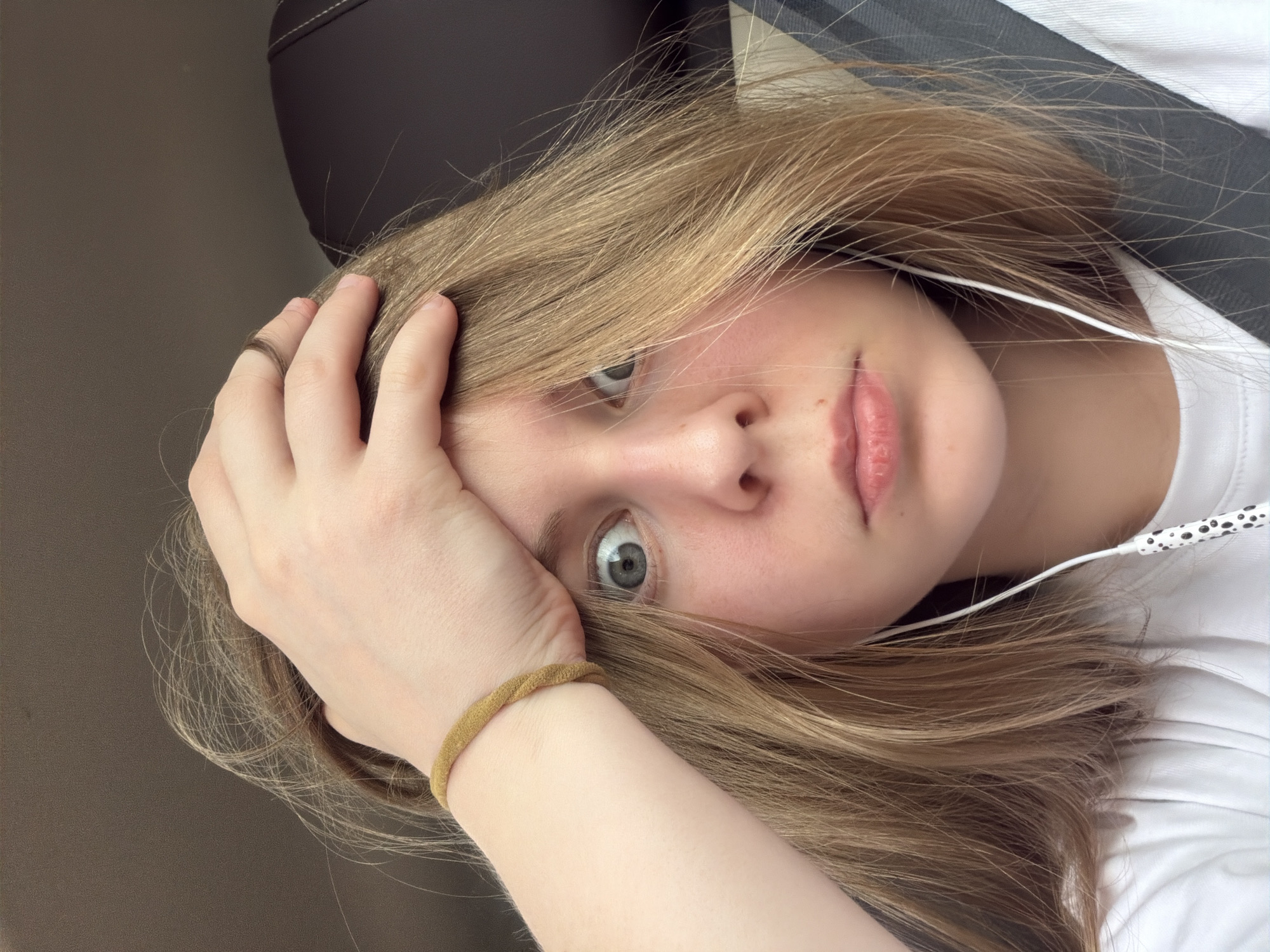 A close-up, selfie-style photograph shows a young person with light brown hair, wide, surprised gray eyes, and fair skin. They are wearing a white t-shirt and have a beaded yellow bracelet on their left wrist. Their right hand is raised, pressing their fingers into their forehead, with their thumb resting on their temple. White earbuds with black polka dots are visible, one dangling near their chin and the other disappearing into their hair. The background is dark, suggesting an indoor setting, possibly a car, with a dark seatbelt visible behind the person. The image is framed from the shoulders up. The location is not specified in the image or its context.