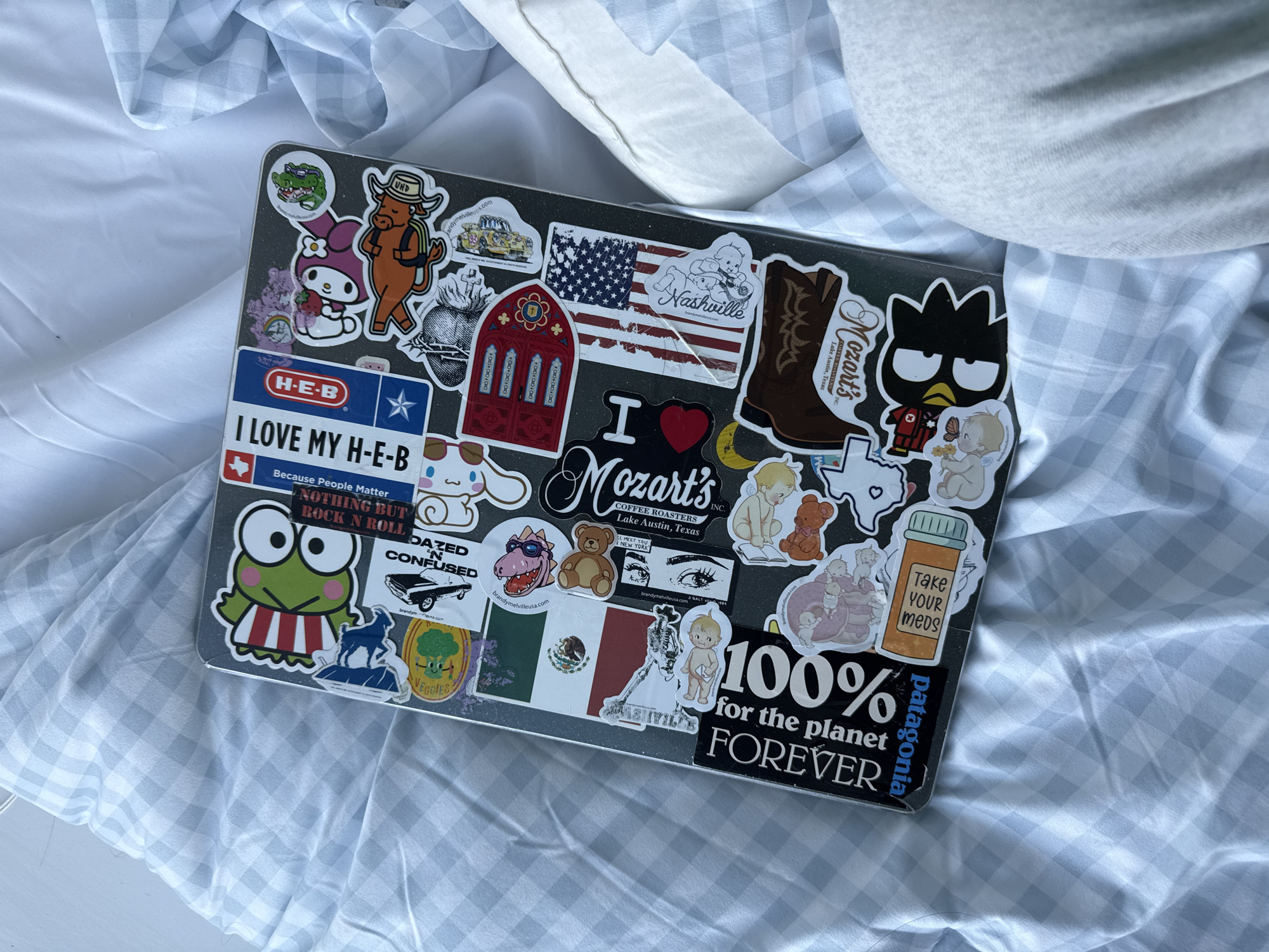 The image shows a laptop covered in stickers on top of a white and blue gingham fabric. The laptop is cluttered with a variety of stickers. Some notable stickers include "H-E-B", "Nashville", "I Heart Mozart's Coffee Roasters Lake Austin, Texas", "Dazed and Confused", "Nothing But Rock N Roll", an American flag, a Mexican flag, and "100% for the planet forever". Mozart's Coffee Roasters is a popular coffee shop located on Lake Austin in Austin, Texas.