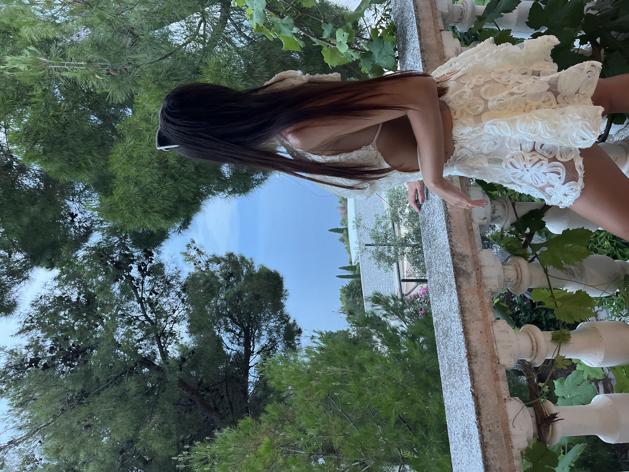 A young woman with long, dark hair and white sunglasses perched on her head stands on a stone balcony overlooking a body of water, likely the Adriatic Sea, and a distant coastline. She is wearing a white, textured dress with cutouts on the sides and a flared, floral-patterned skirt. Lush green pine trees and grapevines frame the scene, suggesting a Mediterranean or coastal environment. The woman is looking out at the view, her right hand resting on the balcony railing. The balcony is supported by ornate white columns and is covered in a fine layer of what appears to be dew or light rain. In the background, there are glimpses of white buildings with grey roofs and scattered cypress trees. The sky is overcast with a soft, diffused light. The location is identified as Šušanj, Montenegro.