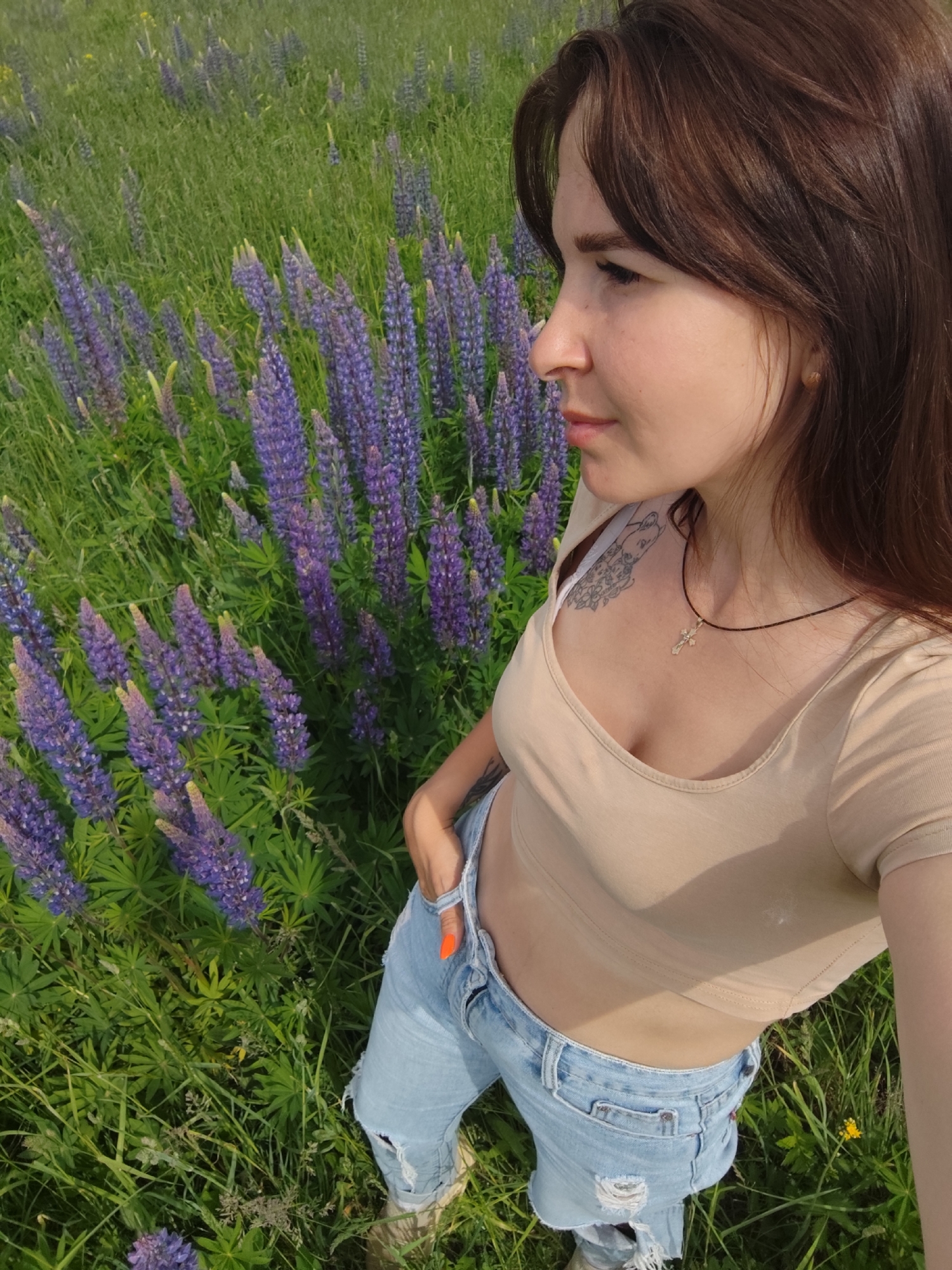 A young woman is captured in a high-angle shot, standing in a field densely populated with tall green grass and numerous purple lupine flowers. She has long, dark brown hair and light skin, and her gaze is directed towards her right (viewer's left) with a neutral expression. She wears a beige short-sleeved crop top and light blue ripped jeans.
Notable details include a dark cord necklace with a silver-colored cross pendant hanging around her neck, resting on her chest. A tattoo is visible on her left shoulder/upper chest area. Her right hand, with bright orange nail polish on her fingernails, is partially tucked into her jeans pocket. The field is lush and green, with the distinctive purple spires of lupines dominating the flora. The bright, clear lighting suggests it is daytime, likely a sunny morning or afternoon. The overall scene conveys a relaxed outdoor setting. The location context for this image is Фоки, Russia. There is no visible text in the image.