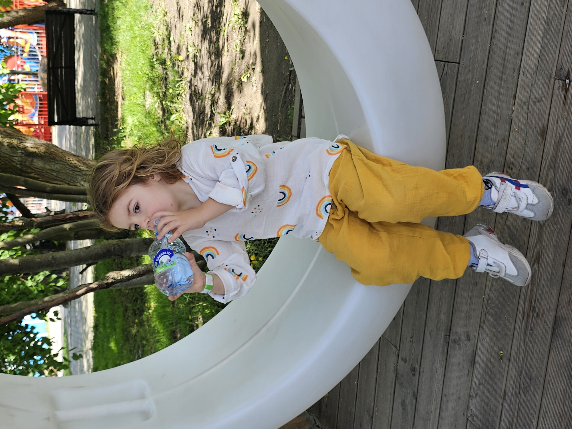 A child, likely between 3 and 5 years old, is drinking water from a plastic bottle. The child has curly blonde hair and is wearing a white shirt with rainbow designs and yellow pants. The child is positioned on a large, white, curved structure, likely part of a playground. The child is also wearing white sneakers with blue and red accents. The structure rests on a wooden deck. In the background, there is a park setting with trees, grass, and a playground structure visible. The photo was taken at Khimki, Russia.