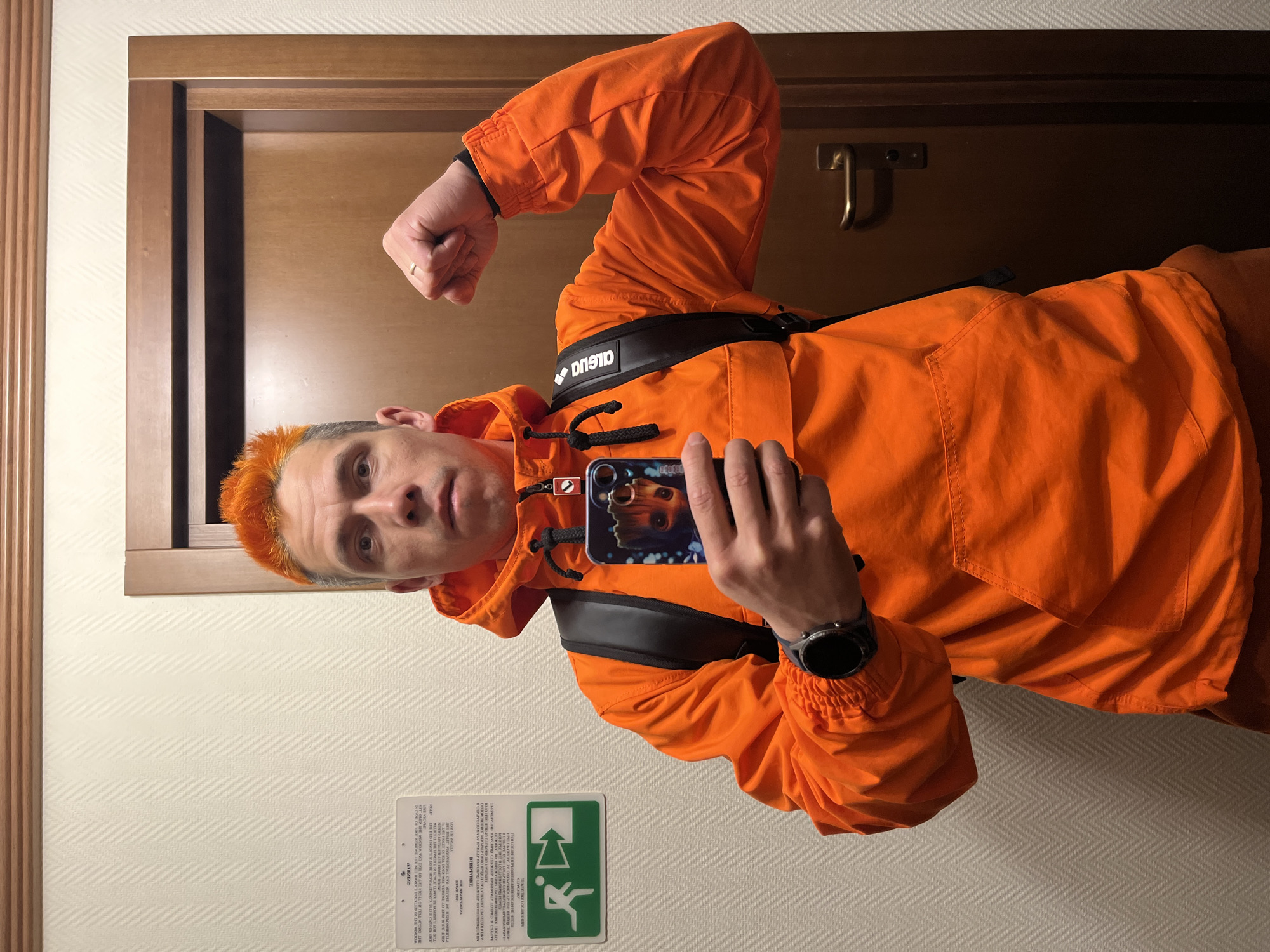 A man with orange hair is taking a selfie indoors. He is wearing an orange jacket with a black chest pack. The background features a door and a wall with an evacuation sign and a framed notice. The location is not identifiable from the image.