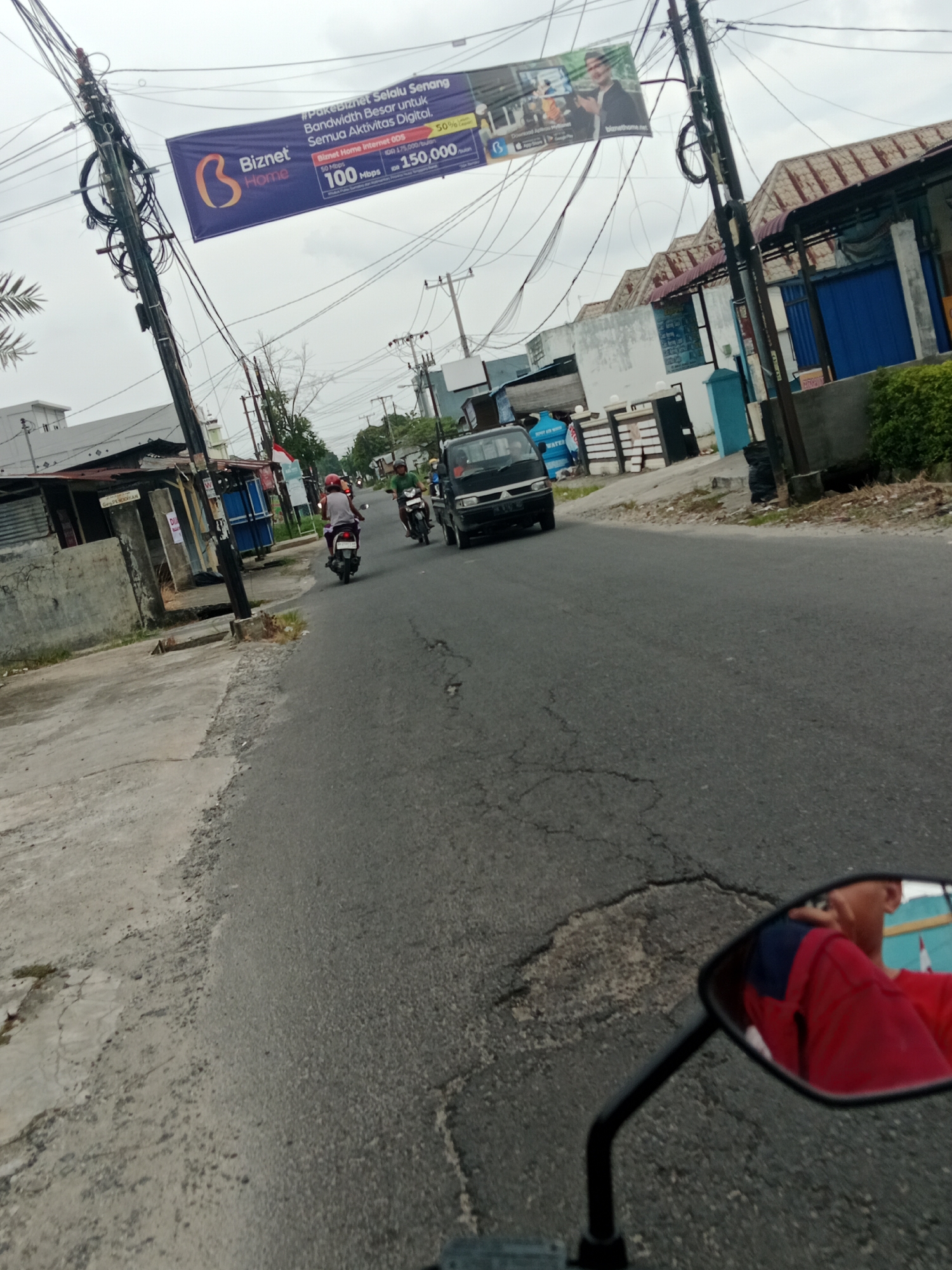A street-level view of a road in the City of Binjai, Indonesia, on an overcast day. The scene is dominated by a large banner advertising "Biznet Home" internet services, suspended across the road from a utility pole. The banner displays text about bandwidth and prices, along with the Biznet logo. Several utility poles with numerous electrical wires are visible, typical of urban infrastructure in Indonesia.
On the road, a black minivan is traveling away from the camera, and two motorcycles are also moving forward. One motorcycle has a rider wearing a red and blue shirt, and another rider is visible further ahead. The road surface is asphalt and shows signs of wear and cracking. To the left of the road, there are buildings with corrugated metal roofs and concrete structures, some of which appear to be shops or houses. Green vegetation is present along the roadside, including some palm trees.
In the foreground, the reflection of the person taking the photo is visible in a motorcycle's side mirror. The reflection shows the person wearing a red shirt. The overall impression is of a typical street scene in a populated area of Indonesia.