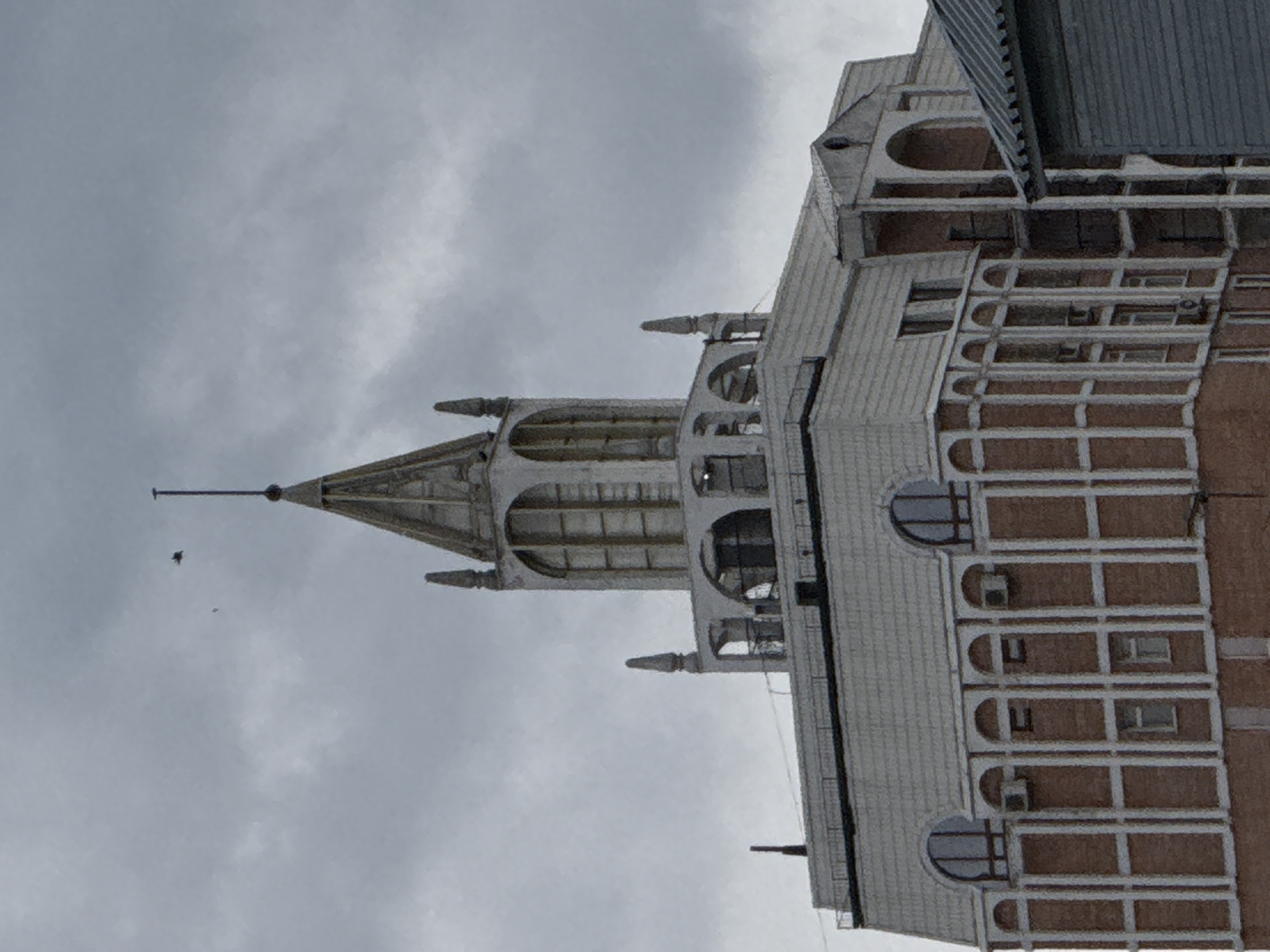 The media file is a vertical, low-angle shot of the upper portion of a tall building, likely a skyscraper or government building, against a cloudy sky. The building features a distinctive architectural style with a pointed spire-like structure at the top, adorned with arched openings and decorative elements. The facade is a mix of light-colored materials, possibly concrete or stone, and darker brickwork. Numerous windows, some round and some rectangular, are visible, along with what appear to be air conditioning units. The sky is overcast and gray, suggesting a cloudy or possibly rainy day, and the lighting is dim, indicating it is likely not midday. Two small, dark shapes are visible in the sky near the spire, which could be birds. The overall scene evokes a sense of urban architecture and a somewhat somber atmosphere due to the weather. The location context suggests this building is in Moscow, Russia, and the architectural style might reflect Soviet or post-Soviet design. No visible text is present in the image.