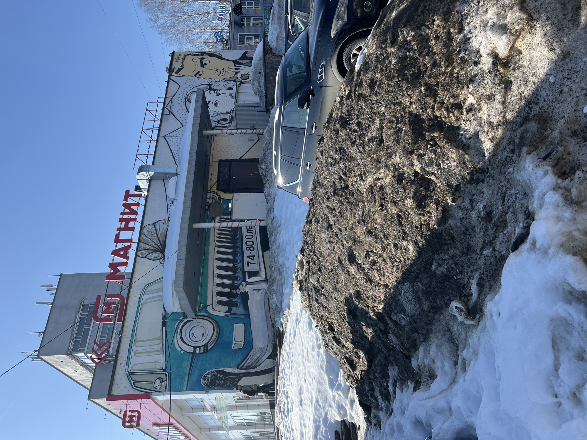The image captures an urban scene in Chaikovsky, Russia, on a clear, sunny day, with conditions indicative of winter or early spring. In the immediate foreground, large, uneven mounds of dirty, melting snow and ice are visible, with embedded litter including a plastic bag. Behind these snow piles, a dark Infiniti QX80 SUV is parked, partially obscured, alongside other vehicles. The main subject is a multi-story building, prominently displaying the red Cyrillic sign "МАГНИТ" (Magnit) across its upper facade. The building's lower section is covered by a detailed mural: a teal vintage car with "74-80 ОЛЕ" written on its front bumper area occupies the left side, while pop art style faces of a woman and a man are depicted on the right. A single individual, dressed in dark attire, walks from left to right along the pavement in front of the building's glass-fronted section and the car mural. The upper portion of the building is a plain gray concrete structure with windows and communication antennas on its flat roof. Bare deciduous trees are visible in the background on the far right.