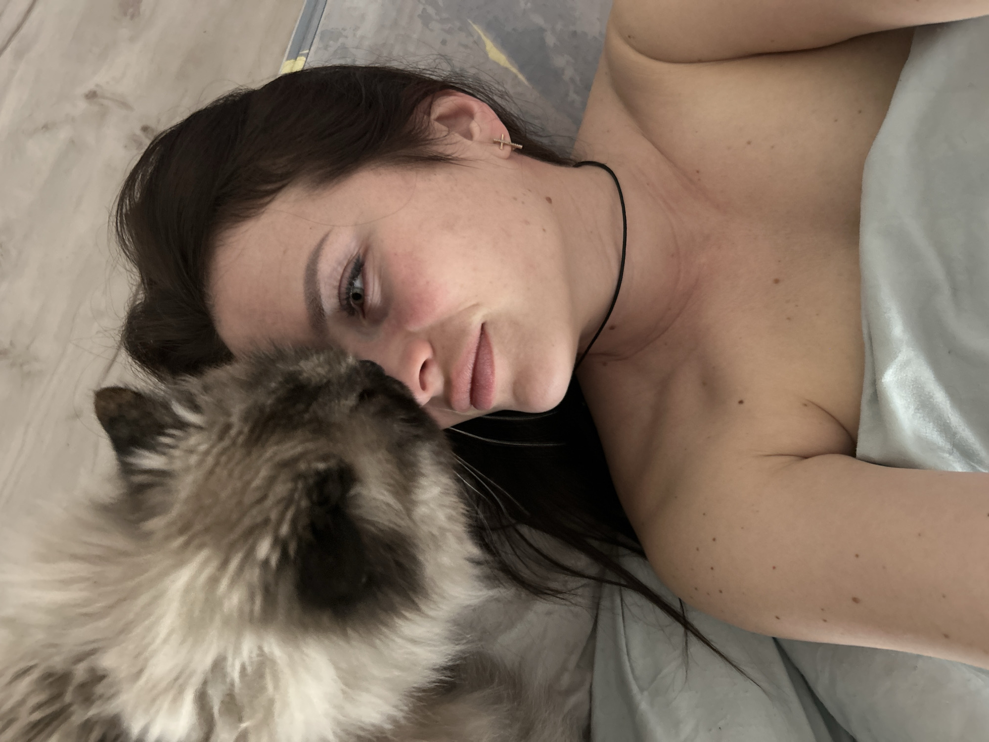A woman with long, dark hair is lying on a bed or surface with a light-colored duvet or sheet. She has a fair complexion, freckles, and is wearing a thin black choker necklace and a cross-shaped earring. Her head is turned towards the left side of the frame, and she appears to be looking at or interacting with a fluffy cat. The cat, a long-haired breed with a mix of dark and light fur, is positioned close to the woman's face, possibly nuzzling or sniffing her. The background includes a light-colored surface that could be a wall or part of the bed frame. There is no information that identifies the location as Rusalevka, Russia.