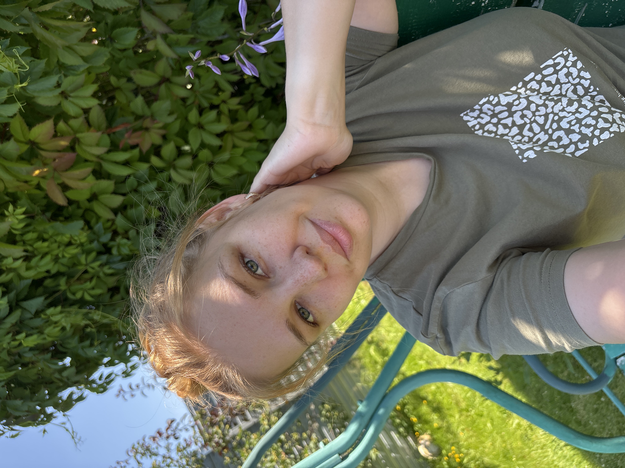 A young woman with light skin and red hair styled in a bun is taking a selfie outdoors. She is wearing a green t-shirt with a white abstract animal print design on it. Her left hand is resting on the back of her neck, and she is wearing a small hoop earring in her left ear. The background consists of lush green foliage and a blue sky. To the left, there is a portion of a teal-colored swing set and a grassy area with yellow flowers. The woman appears to be in a garden or park setting, possibly in the village of Dunilovo, Russia, given the natural environment.