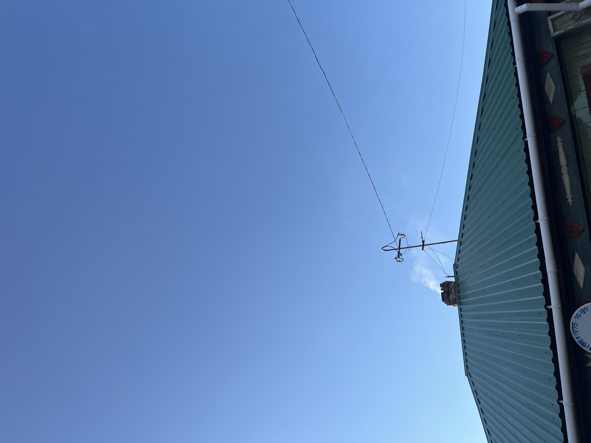 An outdoor, low-angle, full shot captures a clear blue sky above a green metal roof in Бруснятское, Russia. A brick chimney on the roof is emitting a thin plume of white smoke. Above the chimney, a dark, slender electrical wire stretches diagonally across the sky, supported by a utility pole with insulators. The roof is ridged and a white gutter runs along its edge, below which is a dark green facade with decorative elements including red heart shapes and a light-colored wooden spindle. A white circular emblem with black text, partially obscured, is visible on the facade. The lighting suggests it is daytime.
