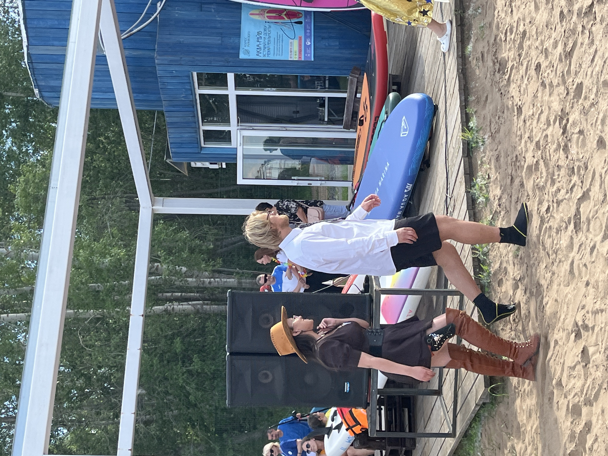 The image captures an outdoor event on a sandy beach, possibly a festival or gathering, with a blue building and trees in the background. A group of people are present, some standing and some seated. In the foreground, two individuals are prominently featured. One person, wearing a white shirt and black shorts, is seated with their legs extended. Beside them, another person is seated, wearing a brown dress and a wide-brimmed hat, with tall brown boots.
Several paddleboards are leaning against a wooden structure. The blue building has windows and a sign with text, though it is difficult to read clearly. One of the paddleboards is blue with "AQUA MARINA" written on it.
The overall atmosphere appears relaxed and casual, suggesting a sunny day, likely daytime. The presence of paddleboards hints at a water-related activity or a venue near a body of water. The sandy beach setting reinforces this.
There is visible text on a sign attached to the blue building, which appears to be in Russian. Additionally, text is present on the blue paddleboard. The image is oriented vertically, with the scene appearing sideways.