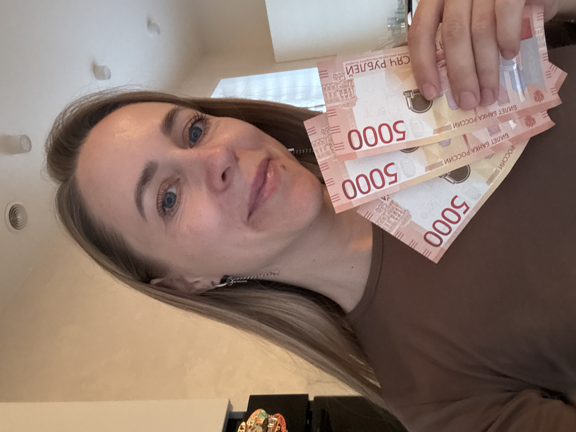 A woman is holding three 5000 Russian ruble bills in her right hand. The bills feature the number "5000" and the text "БИЛЕТ БАНКА РОССИИ" (Bank of Russia Ticket). The woman has blonde hair and blue eyes. The background appears to be a dimly lit room with light-colored walls and a glimpse of a ceiling fixture. There is a dark object on a lower surface, and the woman is wearing a brown top. There is no explicit evidence to confirm the location in the city of Chaikovsky, Russia.