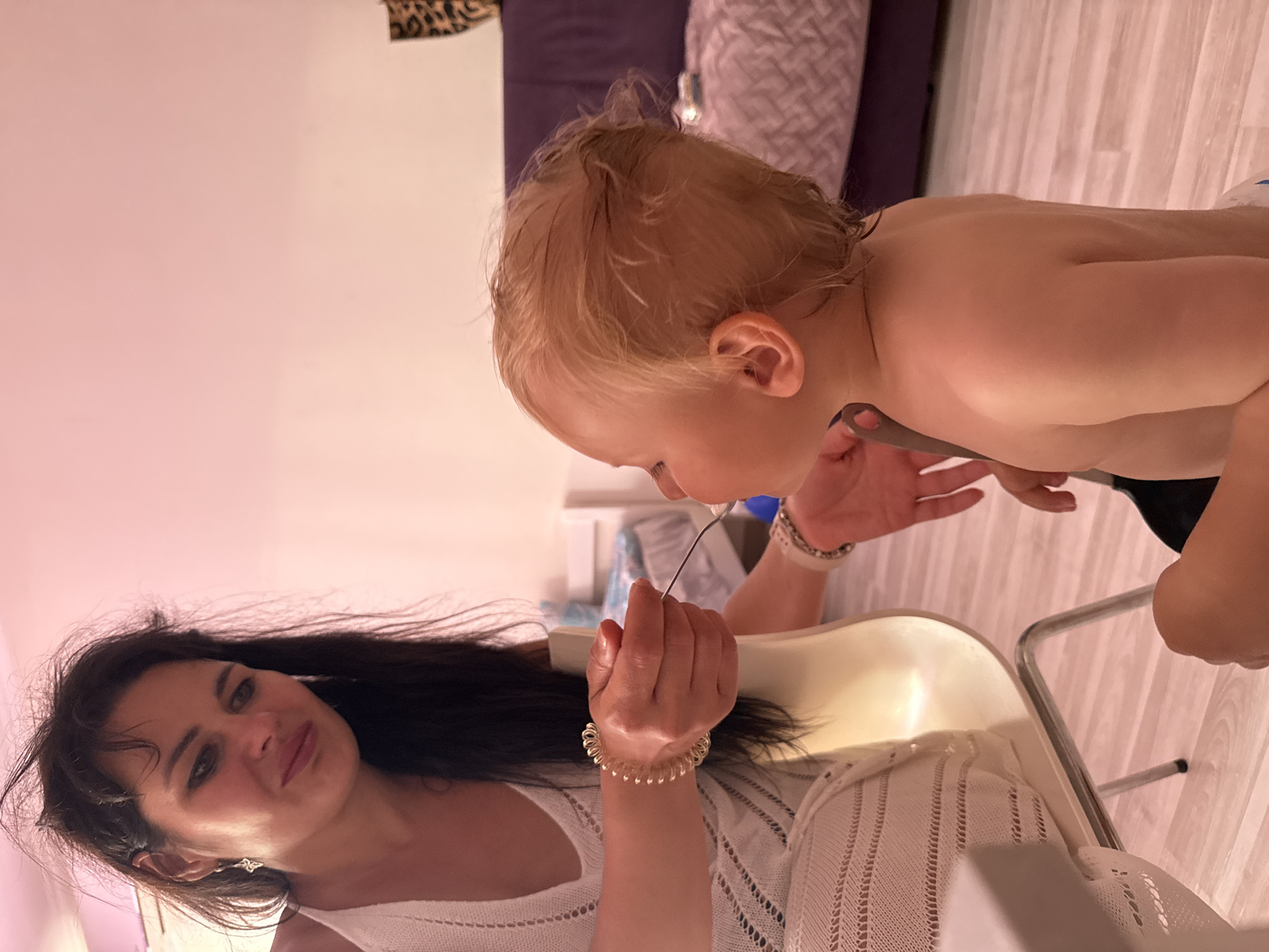 In this media file, a woman with long, dark hair is interacting with a young child who has blonde hair. The woman is looking towards the child with a gentle expression. The child is leaning forward, appearing to be engaged with something the woman is holding. The scene appears to be indoors, with a light-colored wooden floor visible and what looks like furniture in the background. The lighting suggests it might be daytime or evening. The woman is wearing a white top with a textured knit pattern and several bracelets on her wrist. The child is shirtless. The overall mood of the image seems to be one of connection and care between the woman and child.