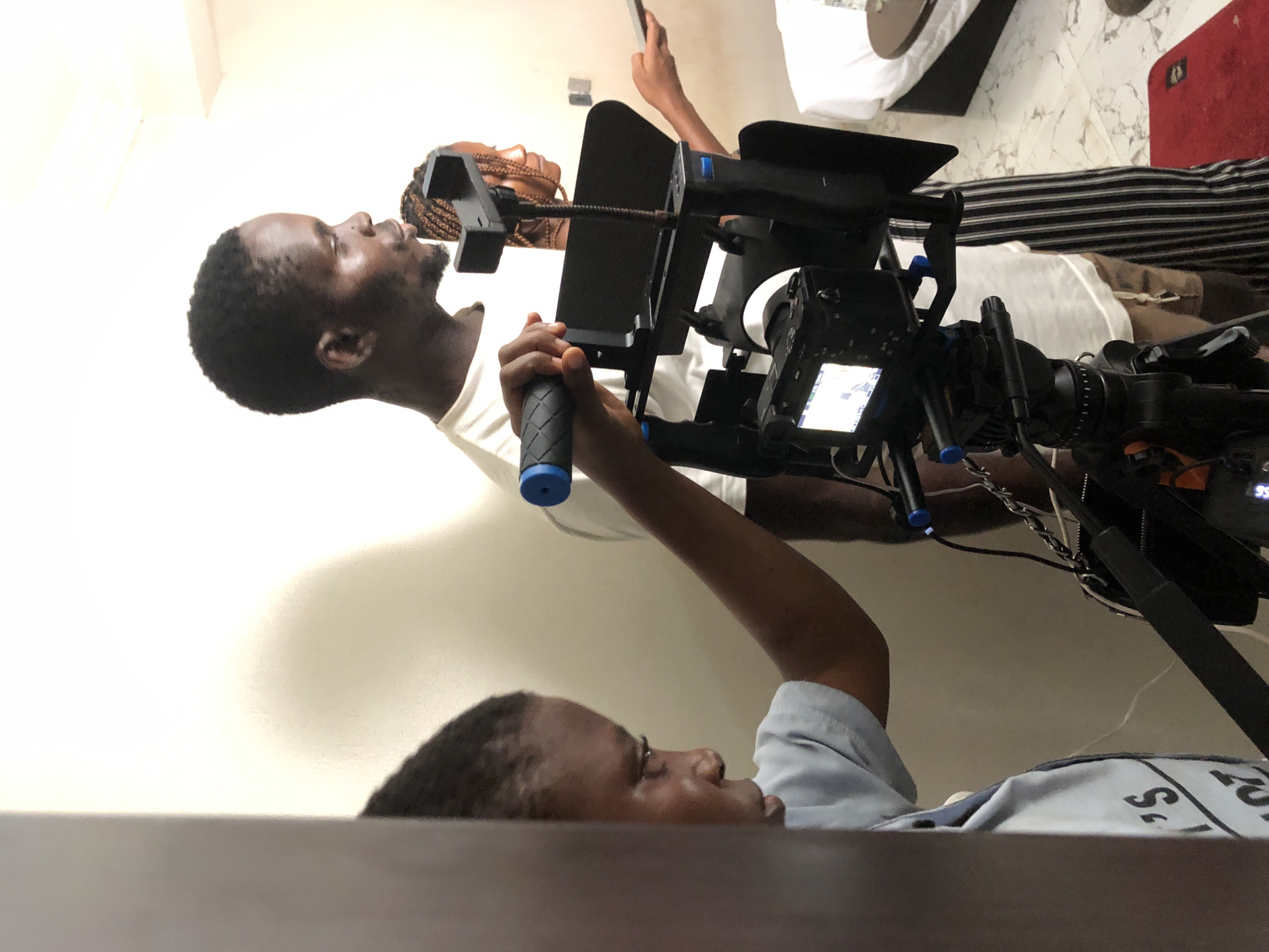 A medium, angled shot depicts a film or video shoot in an interior setting, likely a studio or home location. Two individuals are visible. One man, with dark hair and a dark complexion, is gripping the handles of a camera rig, his face directed towards the subject. The other person is visible in the frame, wearing a white shirt, with braided hair, holding a mobile phone, and facing the camera. A complex camera rig with multiple components is in the center of the frame. The environment consists of a white wall, a bed with a patterned blanket, and part of a table in the foreground.