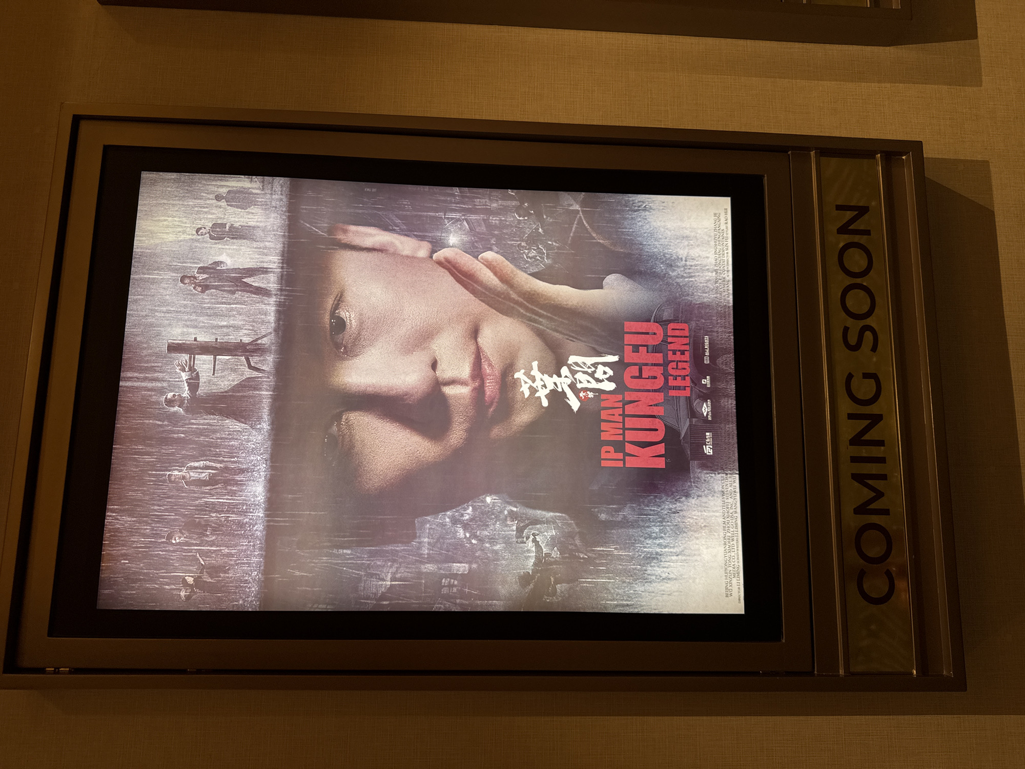 An illuminated movie poster for "IP MAN KUNGFU LEGEND" is displayed in a metal-framed case, likely within a cinema lobby in Denpasar, Indonesia. The poster prominently features a close-up of a person's face, appearing to be a martial artist, with one hand held near their cheek. The overall color palette of the poster is dark and moody, with muted tones and a grunge-like texture, suggesting a serious or action-oriented film. In the background, smaller, silhouetted figures engaged in what appears to be a fight or training sequence are visible, along with what could be rain or atmospheric effects. The Chinese title characters are also present on the poster next to "IP MAN". Below the main title, smaller credits and production information are visible, including names like "BEIJING HUIYONGYUXIAOHU ZHOU XIAOFEIS", "WELL GO USA", and "DIRECTOR LI LIMING". To the right of the movie poster, integrated into the display frame, is a vertical sign with gold lettering that clearly reads "COMING SOON". The surrounding wall has a textured, beige wallpaper. The lighting suggests an indoor setting.