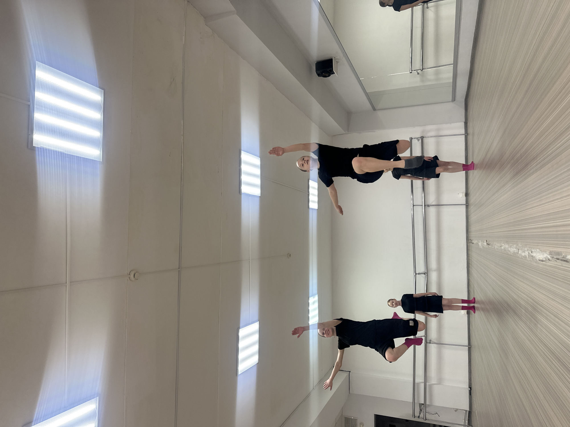 This image depicts an indoor scene, likely a dance or fitness studio located in Votkinsk, Russia. The room is brightly lit by multiple rectangular, grid-patterned LED or fluorescent light fixtures mounted on the white ceiling, which cast strong white light and visible rays onto the surroundings. The floor has a light-colored, wood-like texture with fine horizontal lines. One entire wall is covered by a large mirror, beneath which are two parallel metal barres, characteristic of a ballet or dance studio.
Two individuals are present in the studio, both dressed in dark athletic wear. In the foreground, a young person performs a dynamic balance pose, leaning forward with their torso parallel to the ground, one leg extended backward, and arms outstretched to the sides. They are wearing a black t-shirt, black shorts, and a prominent bright pink sock on their extended foot. Their reflection is clearly visible in the large mirror, appearing as if in a complementary, inverted position.
Further back in the room, a second individual stands upright, also looking towards the front. This person wears a black tunic or short dress over black shorts and distinct bright pink socks. Their reflection is also visible in the mirror, positioned closer to the reflection of the first person.
Additional details include a small, black speaker mounted high on the wall near the ceiling, and a circular smoke detector or similar device affixed to the white ceiling. The overall setting is clean, functional, and well-lit, designed for physical activity. There is no visible text in the image.