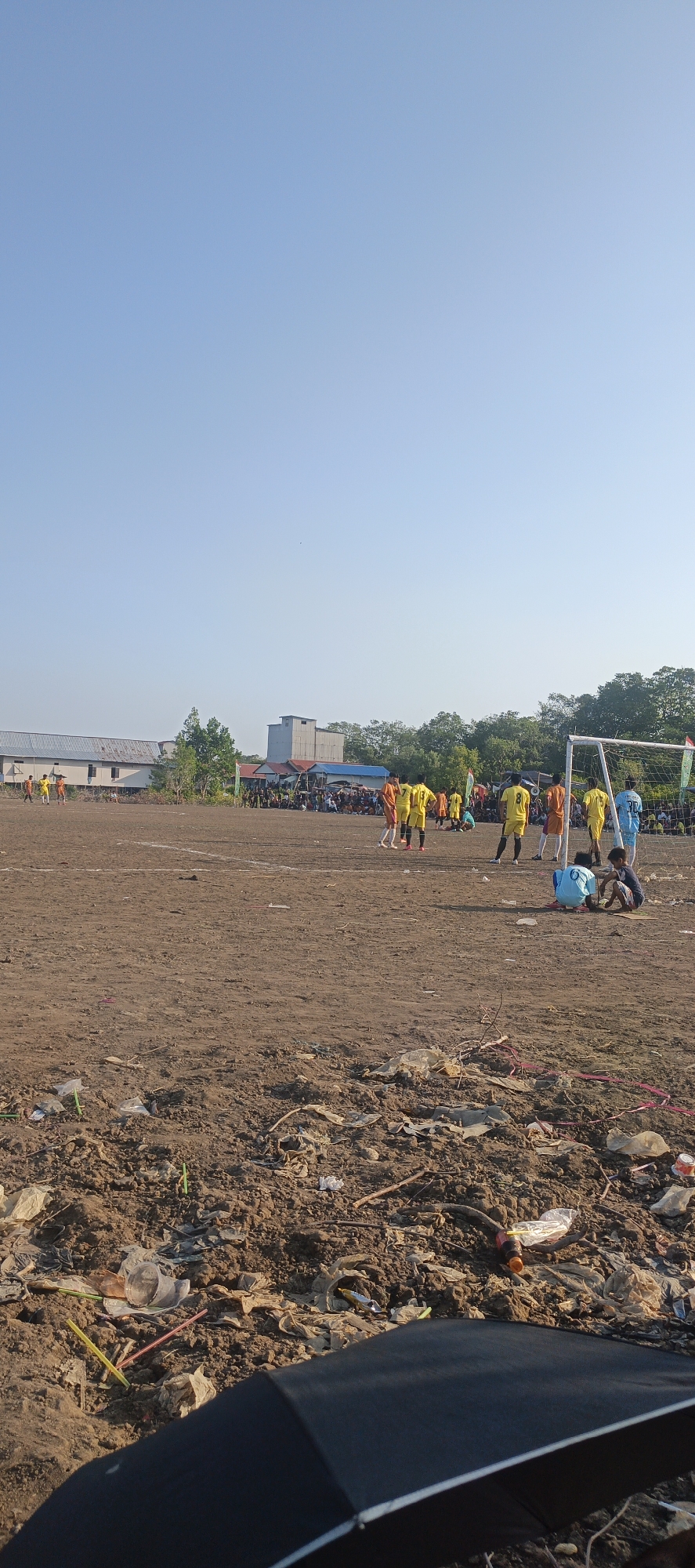 In Lori, Indonesia, a soccer game is underway on a dirt field under a clear blue sky. The field is marked with white lines and shows signs of wear and tear, with patches of exposed earth. Spectators are gathered on the sidelines, some standing and others sitting. Two teams, distinguishable by their yellow and orange jerseys, are engaged in play. A soccer goal stands on the right side of the frame. The foreground of the image is dominated by a pile of debris, including plastic bags, bottles, and straws, indicating a lack of proper waste disposal. A partially opened black umbrella is visible in the bottom right corner of the frame, suggesting someone is using it for shade. In the background, buildings and trees form the backdrop to the soccer field.