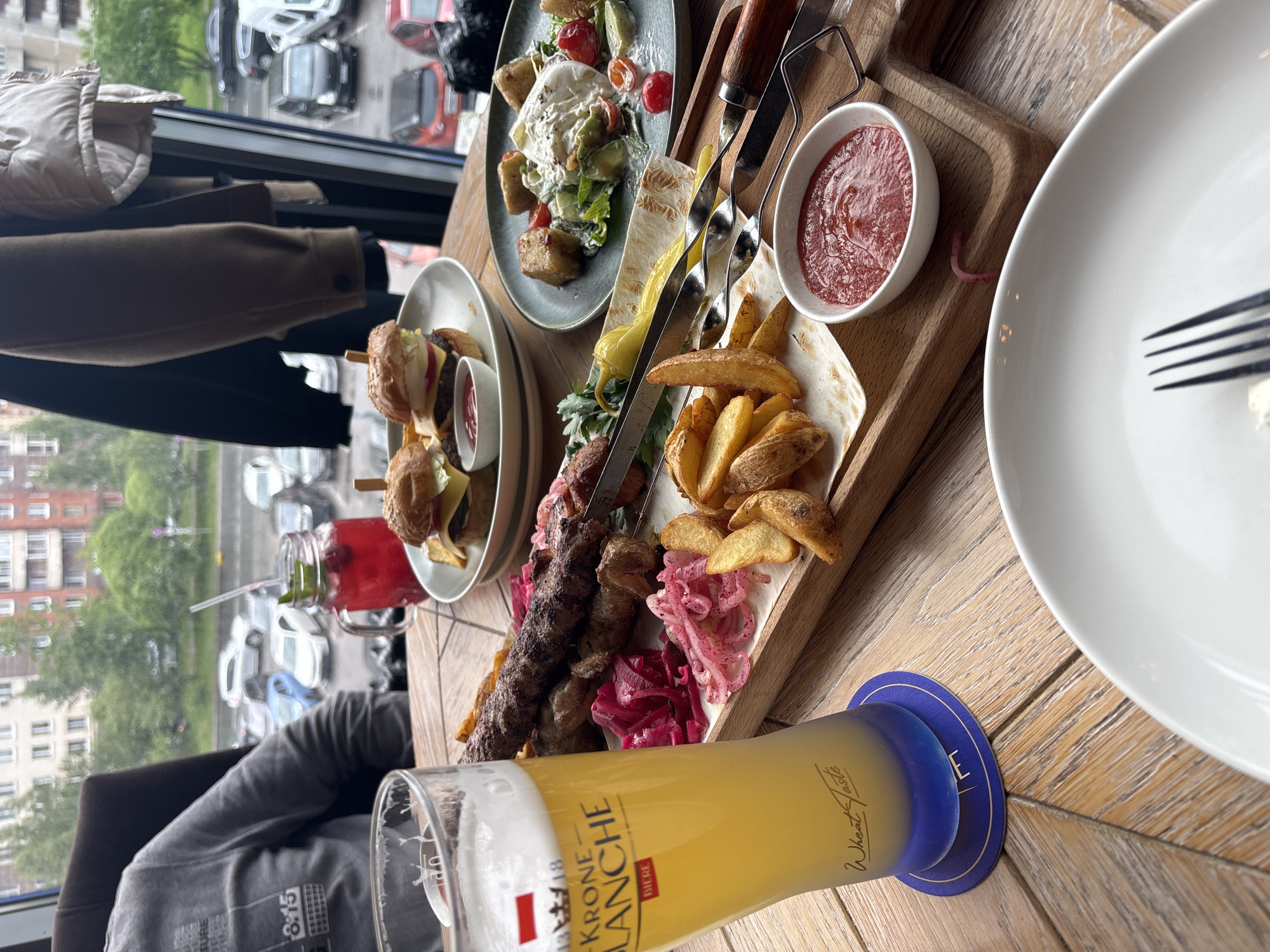 The image captures a restaurant scene in Saint Petersburg, Russia, featuring a wooden table laden with an assortment of food and drinks. Dominating the foreground is a tall glass of light-colored beer, partially obscuring a wooden platter arranged with shish kebabs on skewers, accompanied by fried potato wedges, lavash bread, pickled red cabbage, and a small bowl of red sauce. Additional dishes include a salad with white cheese and cherry tomatoes, as well as mini-burgers served with dipping sauce. A white plate with a fork sits to the right. In the background, through a window, there is a view of a city street with parked cars and buildings. A person wearing a grey sweatshirt is partially visible in the lower-left corner of the frame. A dark-colored jacket hangs near the window.