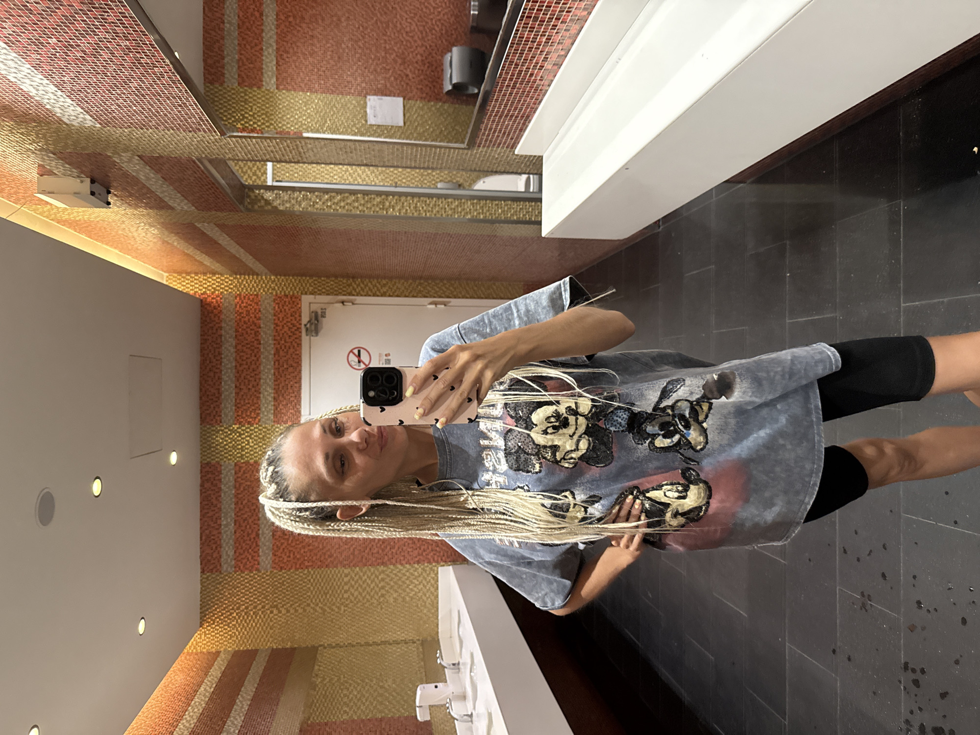 A woman with blonde braided hair takes a mirror selfie in a modern bathroom. She is wearing a grey t-shirt with a Mickey Mouse graphic and black biker shorts. The bathroom features patterned orange and yellow tiled walls, white sinks, and dark tiled floors. A toilet and a paper towel dispenser are visible in the reflection of the mirror. The lighting is bright, likely from overhead fixtures. There is no indication of the city of Yaroslavl, Russia in the image.