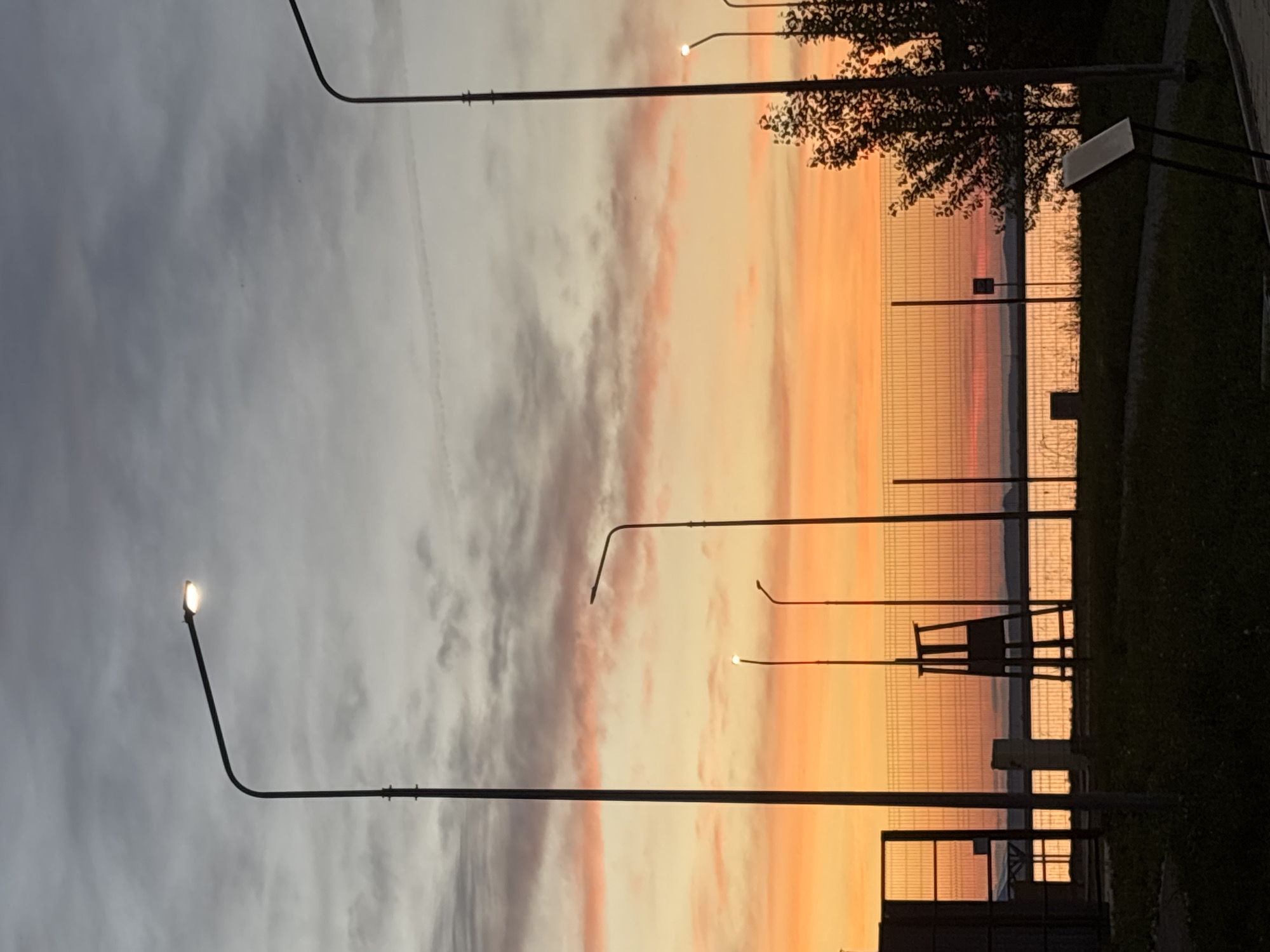 The image depicts a sunset over the Kama River in Chaikovsky, Russia. The sky is a vibrant gradient of orange, yellow, and pink, with scattered gray clouds. Several streetlights are visible, some illuminated, casting a warm glow. In the foreground, there is a grassy area with a paved path and a few dark-colored signs. A chain-link fence runs across the middle ground, separating the paved area from the river. Behind the fence, a lifeguard chair or observation post stands silhouetted against the bright sky. Trees with dark foliage are present on the right side of the frame. The river is calm and reflects the colors of the sunset. The overall atmosphere is serene and tranquil.