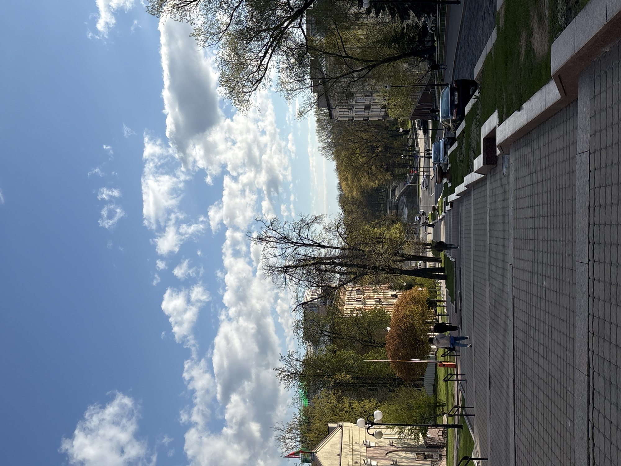 The image depicts a view from an elevated position, likely steps or a platform, overlooking a cityscape in Minsk, Belarus. The foreground consists of a paved area with steps leading down to a street lined with trees. A few people are walking along the paved area and the street. Beyond the trees, buildings are visible. Cars are parked or driving along the street. The sky is blue with scattered clouds.