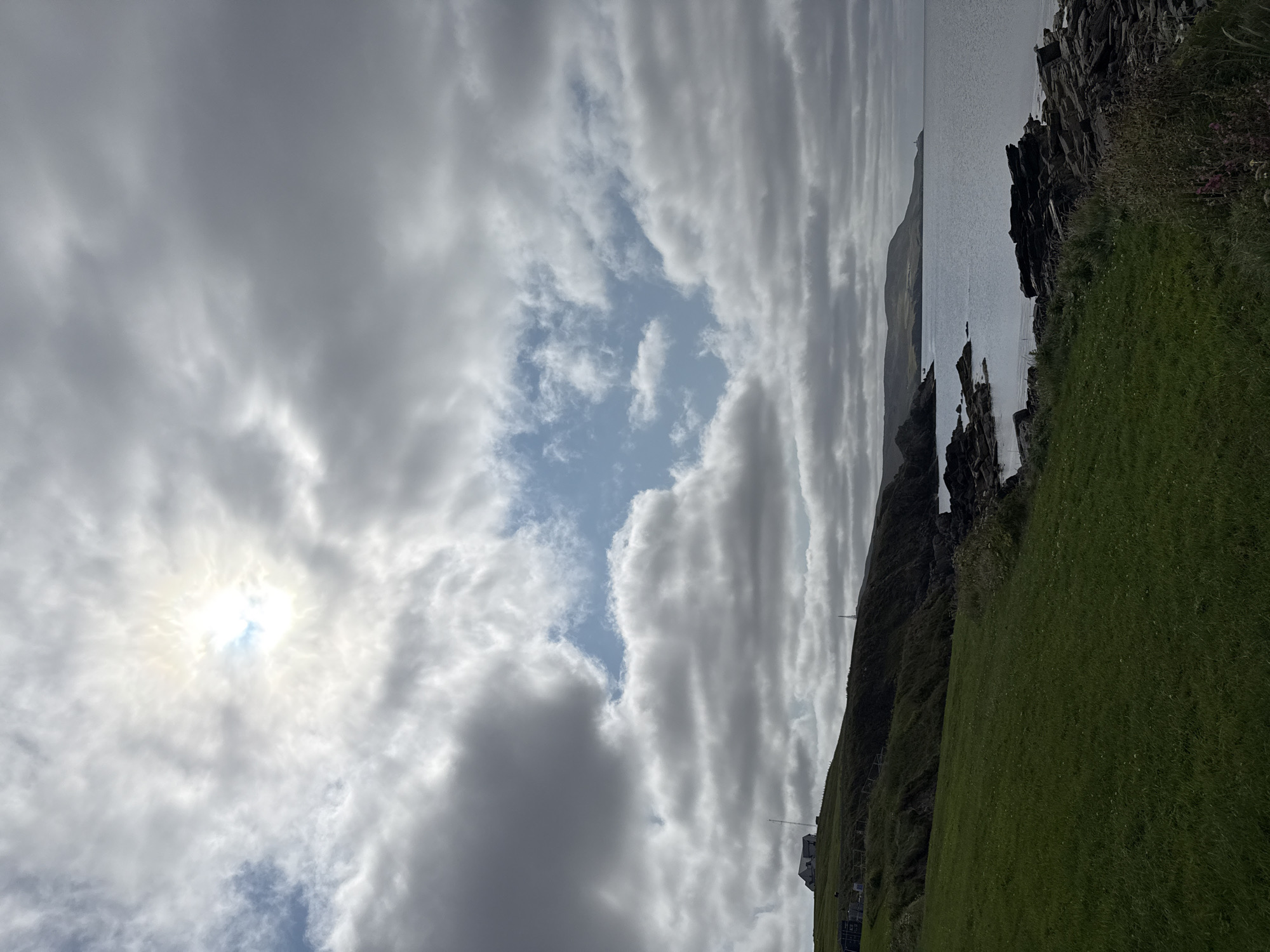 This image depicts a cloudy coastal landscape. The foreground is dominated by a lush green grassy field sloping down towards a rocky shoreline. The sea is calm and gray, meeting a distant, hazy coastline with rolling hills. A prominent feature on the hill to the left is a white house. The sky is filled with a mix of dark and light gray clouds, with a bright patch of sun breaking through in the upper center, creating a halo effect. The overall impression is one of a serene, possibly windswept, coastal environment. The location is identified as Lerwick, United Kingdom. There are no people or active subjects visible in the scene. The environment appears natural and undeveloped, with the exception of the single dwelling on the hillside.