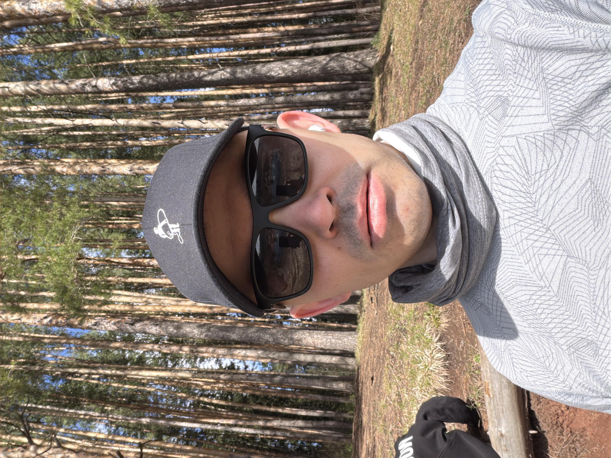 A selfie photograph depicts a man in front of a dense forest with tall, straight trees. The man is wearing a gray baseball cap with a white graphic on the front, dark sunglasses, and a gray patterned top with a gray neck gaiter. He appears to be standing in a forest, possibly in Izhevsk, Russia, given the presence of dense pine forests in the region. The ground is covered with brown pine needles and some grass.
