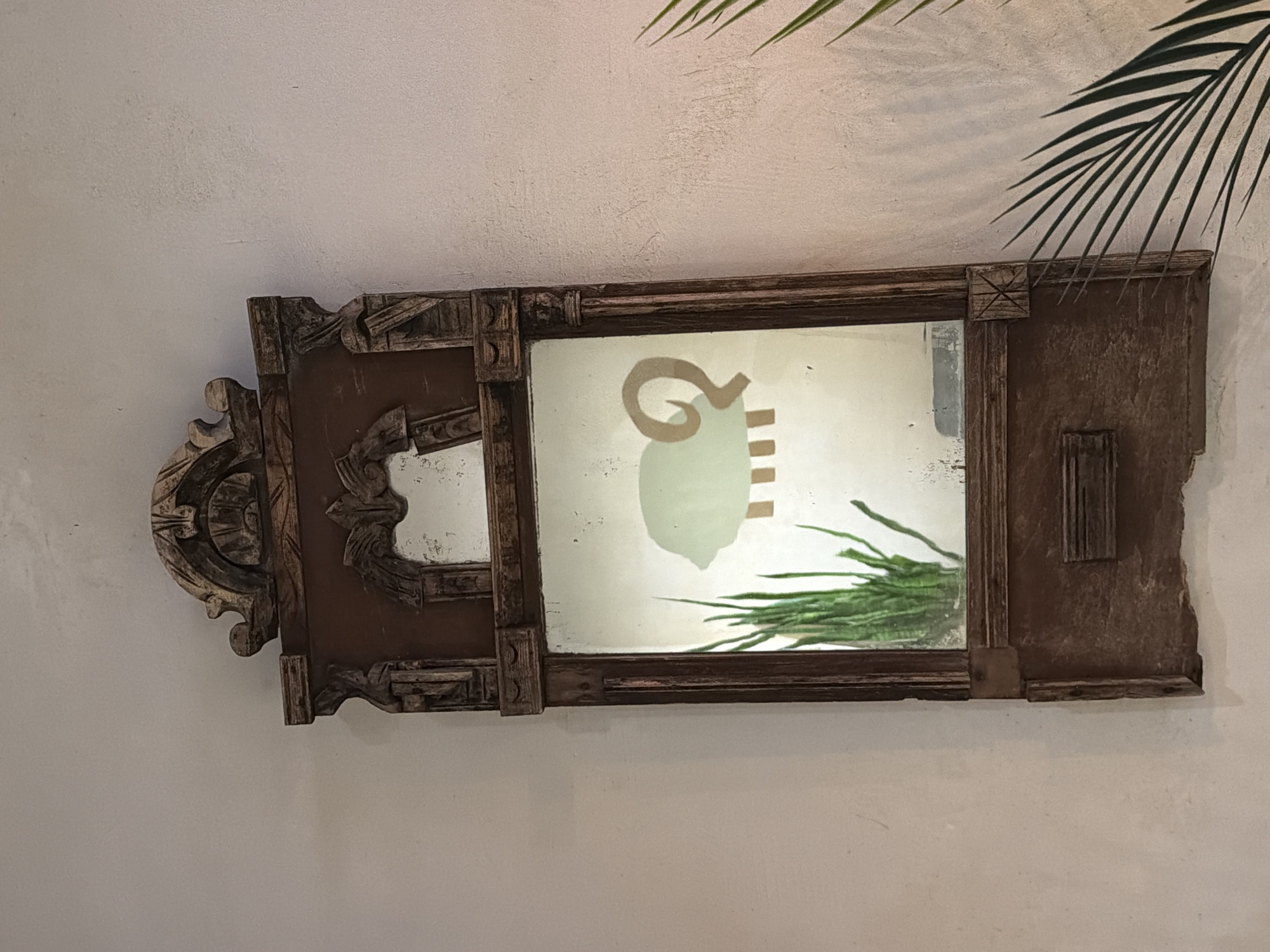 An ornate, rectangular mirror with a dark, carved wooden frame is mounted on a plain, light-colored wall. The mirror is oriented horizontally, with its left side featuring intricate, scrolled carvings and decorative elements, while the right side has a simpler, more rustic appearance with a roughly cut edge and some vertical detailing. The wood of the frame appears aged and has a textured, possibly distressed, finish.
Reflected in the mirror glass, which shows some signs of age or wear, is a stylized, light green silhouette of an animal head, resembling a ram or goat, complete with a prominent curled horn. Below this animal motif, several short, light brown horizontal lines are visible. To the bottom right within the reflection, a vibrant green plant, possibly the fronds of a fern or palm, is reflected. Overhead, outside the mirror's frame and extending from the top right corner of the image, is a real palm frond casting a soft shadow on the wall.
The wall itself is a uniform, light off-white or cream color with a slightly textured finish. The overall scene suggests an indoor setting, possibly a decorative element in a home, a restaurant, or a hotel, with a rustic yet elegant aesthetic. There are no people present, and no specific activity or event is taking place. The lighting is soft and ambient, making the time of day indeterminate. No text is visible in the image.
